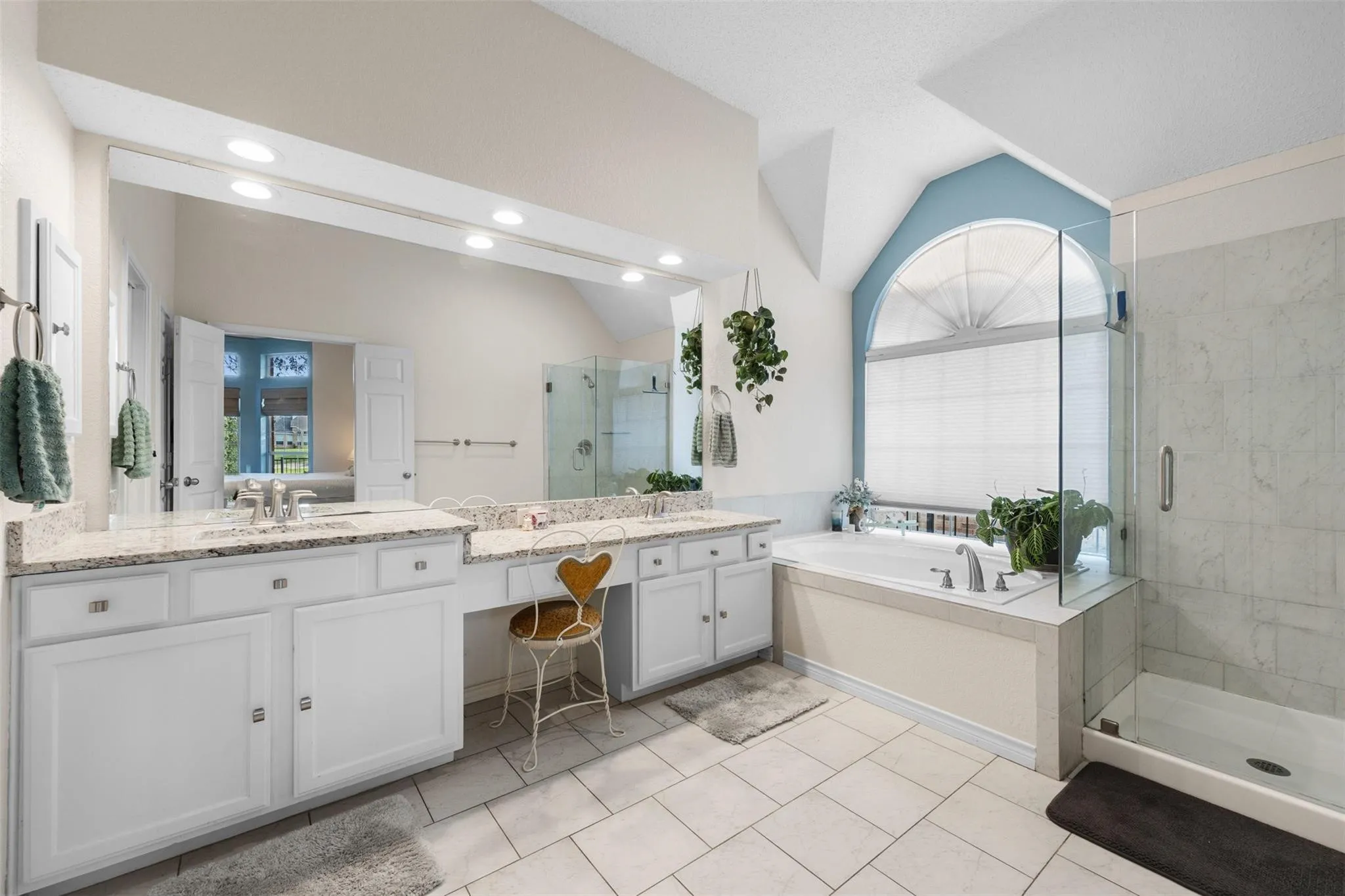 Dual vanity with light-toned countertops and undermount sinks, glass-enclosed shower, built-in soaking tub, and arched window