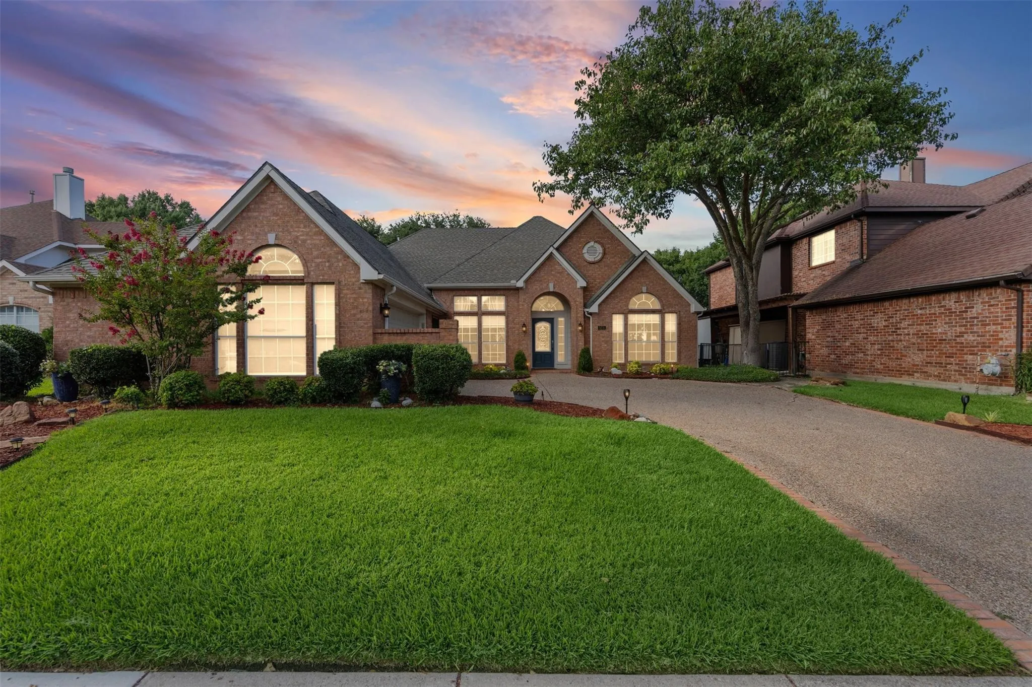 Brick exterior with arched windows, mature landscaping, a paved driveway, and an attached garage