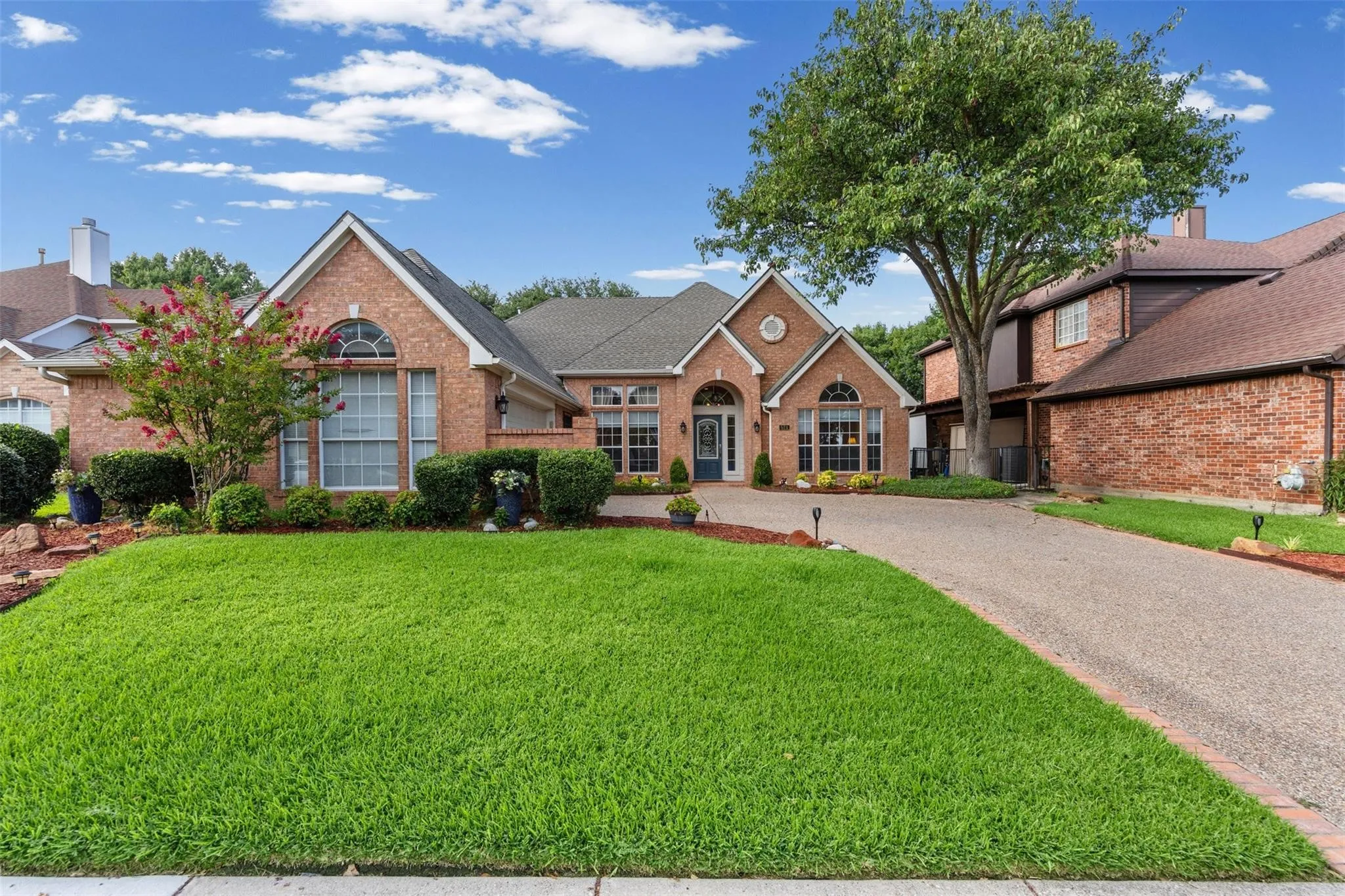 Brick Colonial home featuring a manicured front lawn, curved driveway, arched entryway with transom window, and mature landscaping including flowering shrubs and established trees