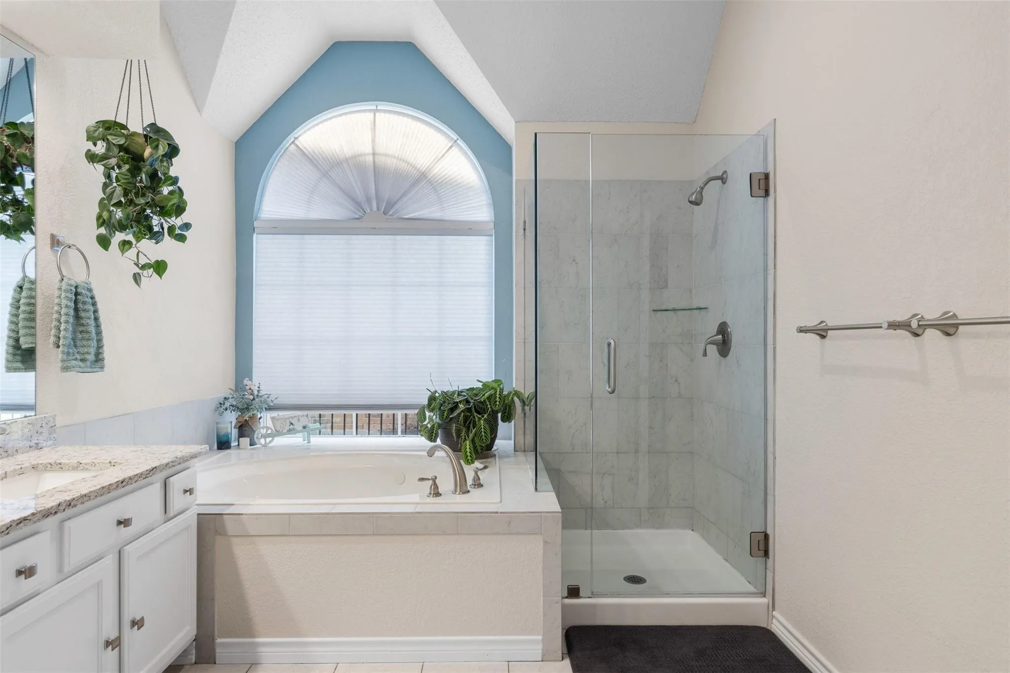 Built-in soaking tub, arched window with privacy blinds, glass-enclosed shower, white vanity with granite-style countertop, and tile flooring