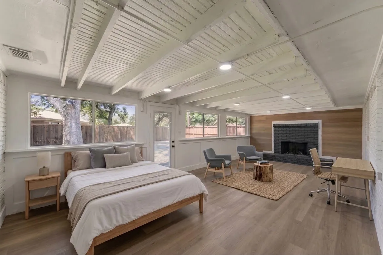 Vaulted ceiling with exposed beams, wood-finish flooring, multiple windows, black brick fireplace with wood paneling surround, and recessed lighting