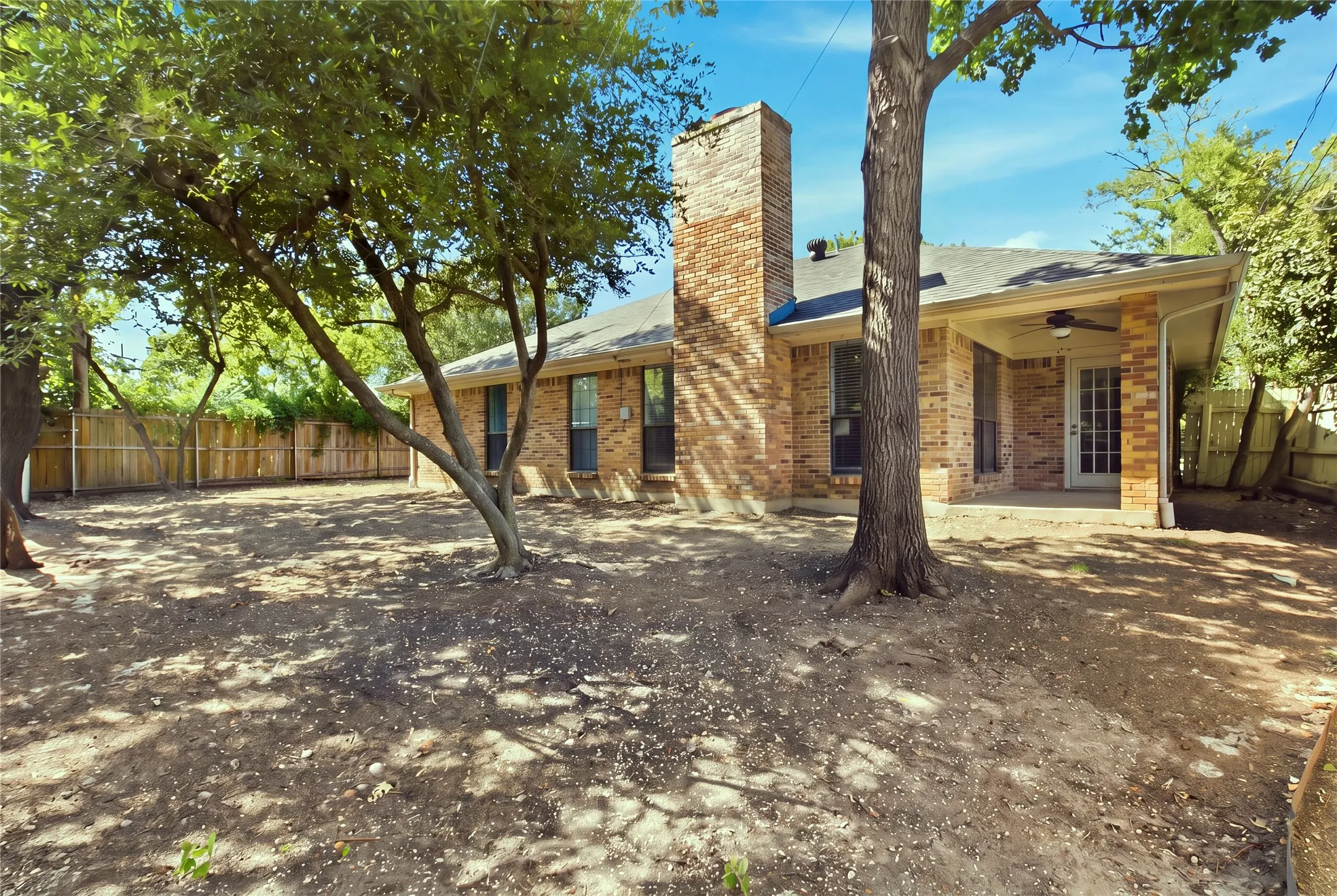 Brick exterior with a prominent brick chimney, mature trees, a covered patio with a ceiling fan, and a wood privacy fence