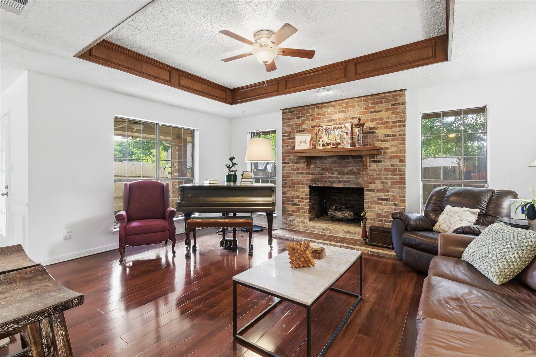 Wood-finish flooring, brick fireplace with mantel, tray ceiling with wood accents, and ceiling fan with light fixture