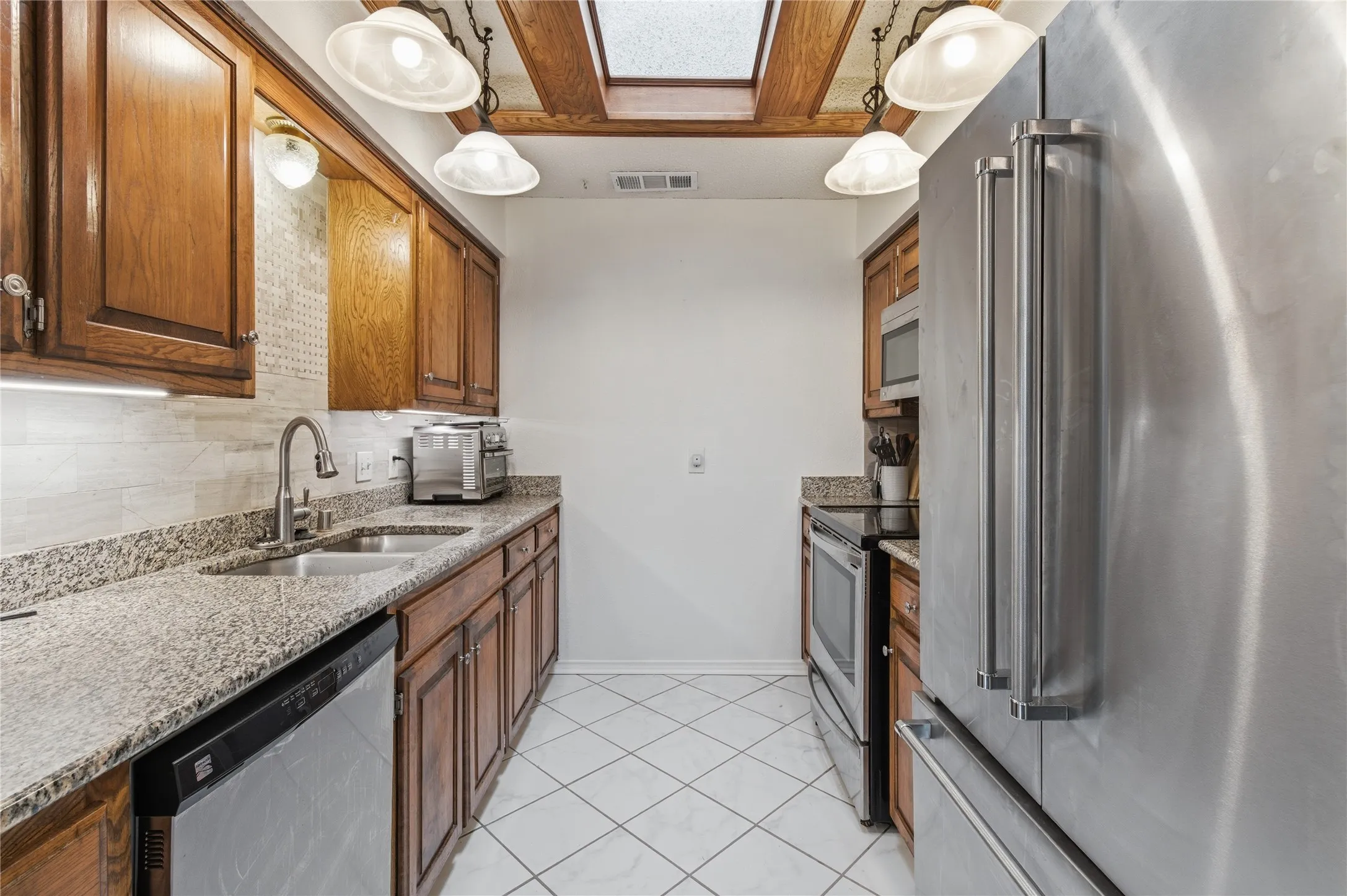 Galley kitchen featuring wood cabinetry, granite countertops, stainless steel appliances, a double basin sink, and a skylight