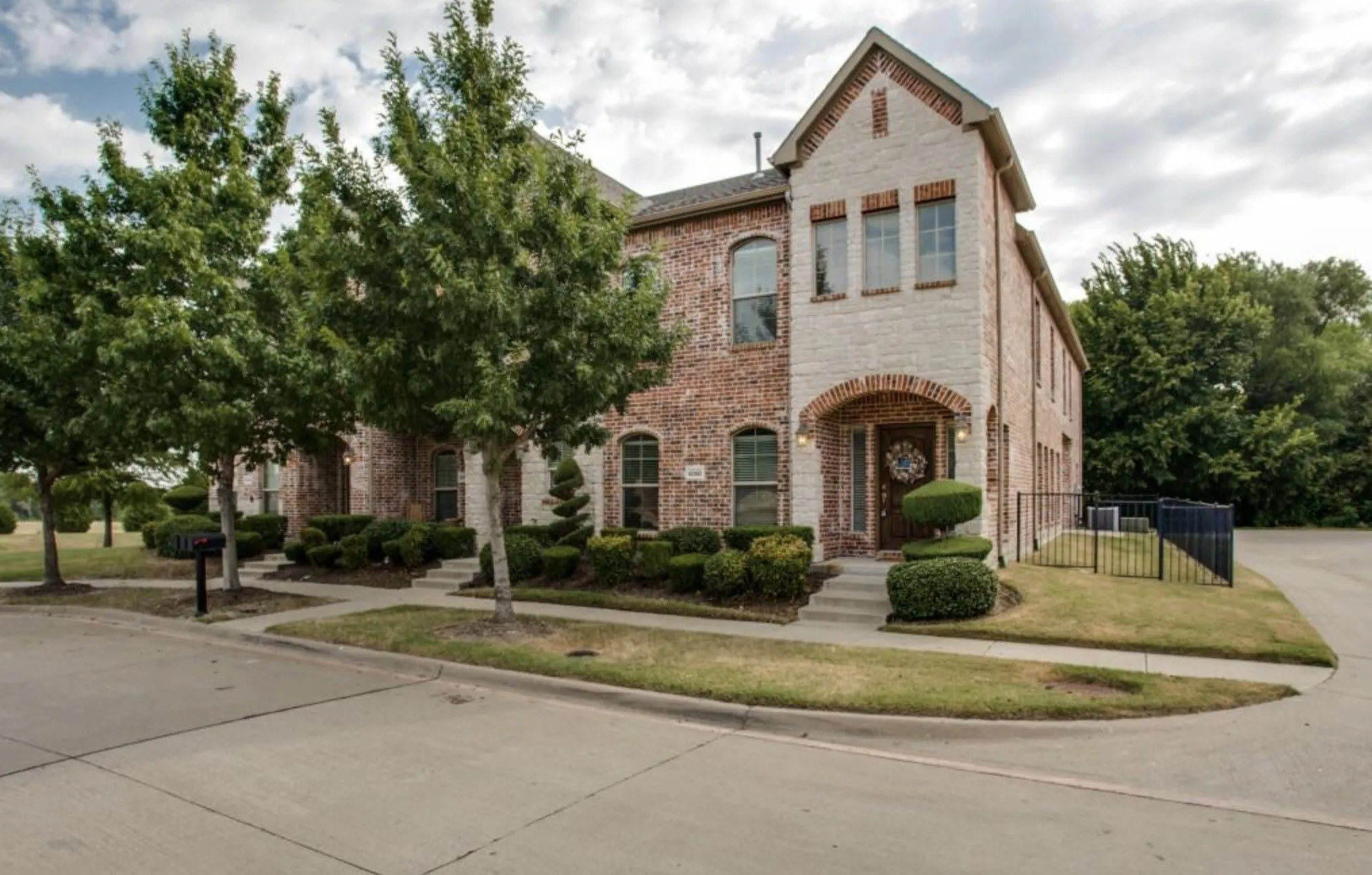 Brick and painted brick exterior with arched entryway, mature trees, and manicured shrubbery