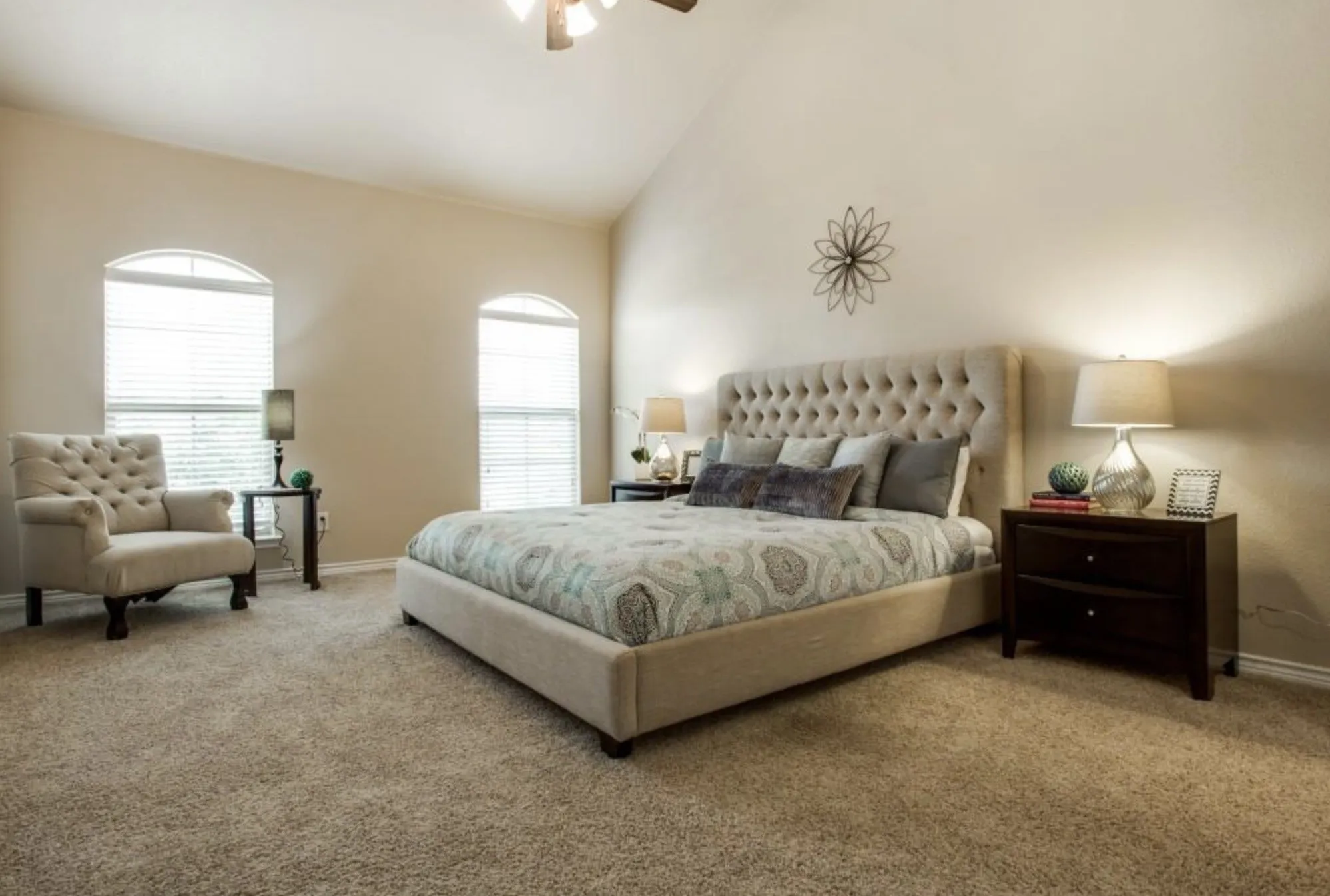 High vaulted ceiling, dual arched windows with blinds, plush carpet flooring, ceiling fan with integrated lighting, and neutral wall tones