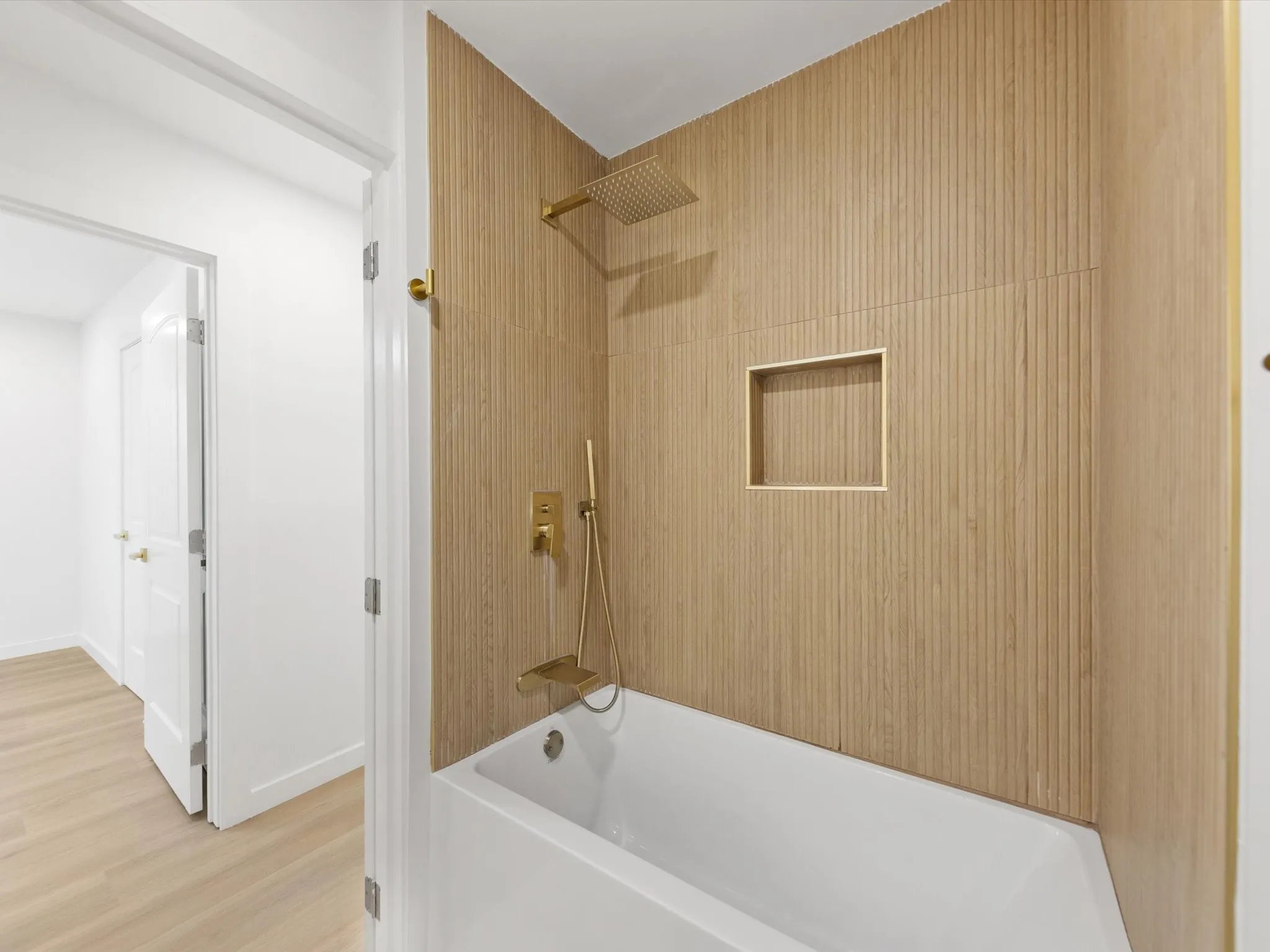Bathtub with ridged wood-finish wall paneling, built-in shower niche, and brass fixtures