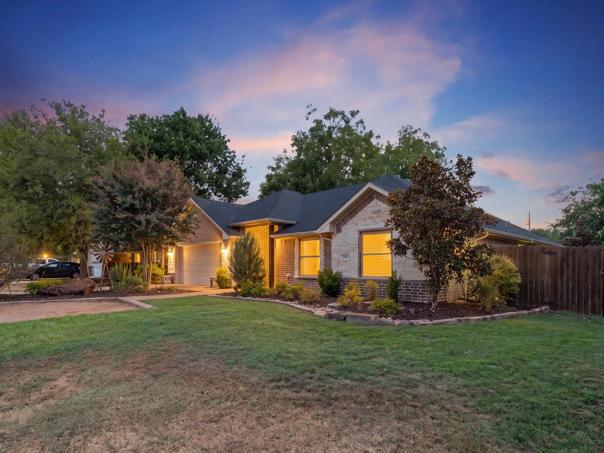 Brick and stone facade, attached two-car garage, dark-pitched roof, established landscaping, and a wooden privacy fence