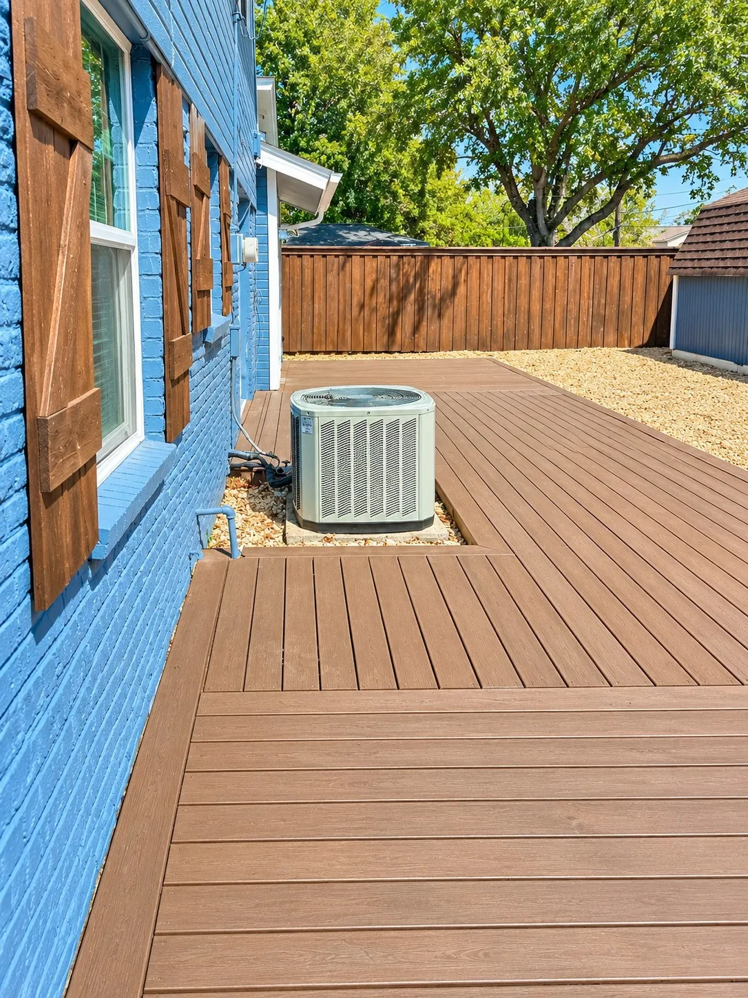Expansive composite decking with an integrated air conditioning unit, brick exterior with wood shutters, and a wood privacy fence