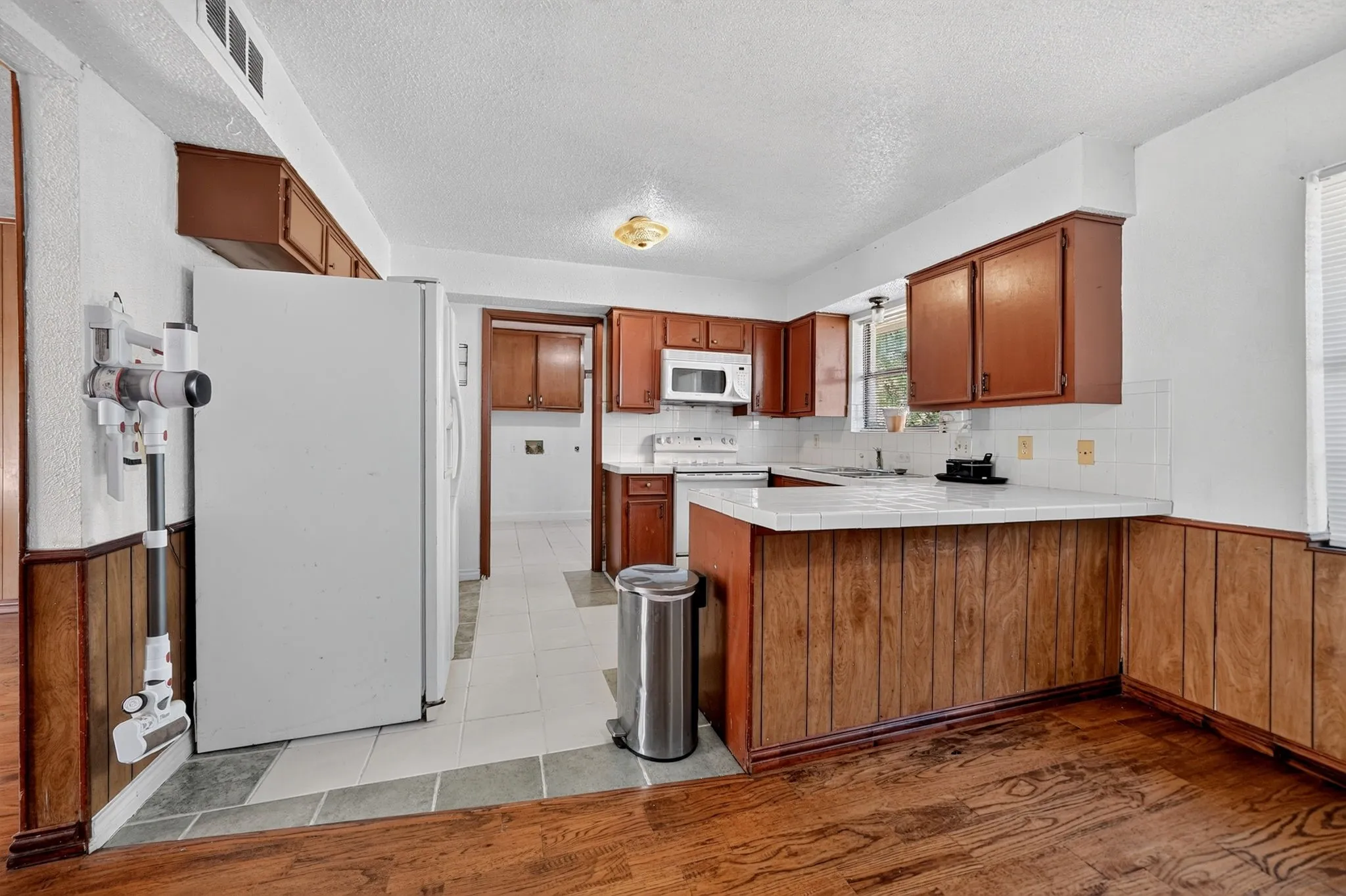 Wood-finish flooring, wood paneling, white ceramic tile countertops, white tile backsplash, and brown cabinetry