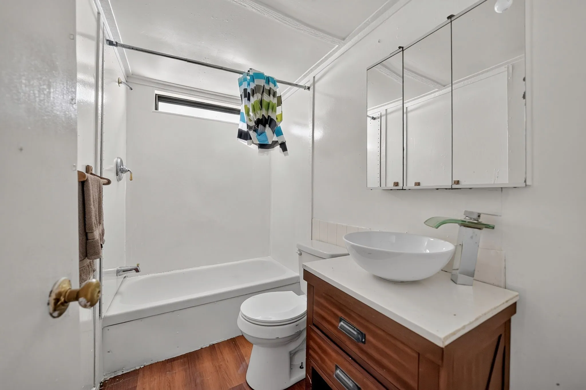 Bathtub and shower combination, dark wood-finish flooring, white vanity with countertop, vessel sink, and mirrored medicine cabinet