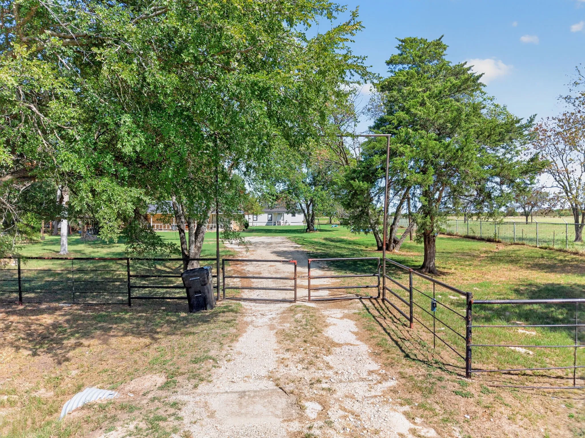 Gravel driveway with metal gates and fencing, surrounded by mature trees and expansive grassed areas