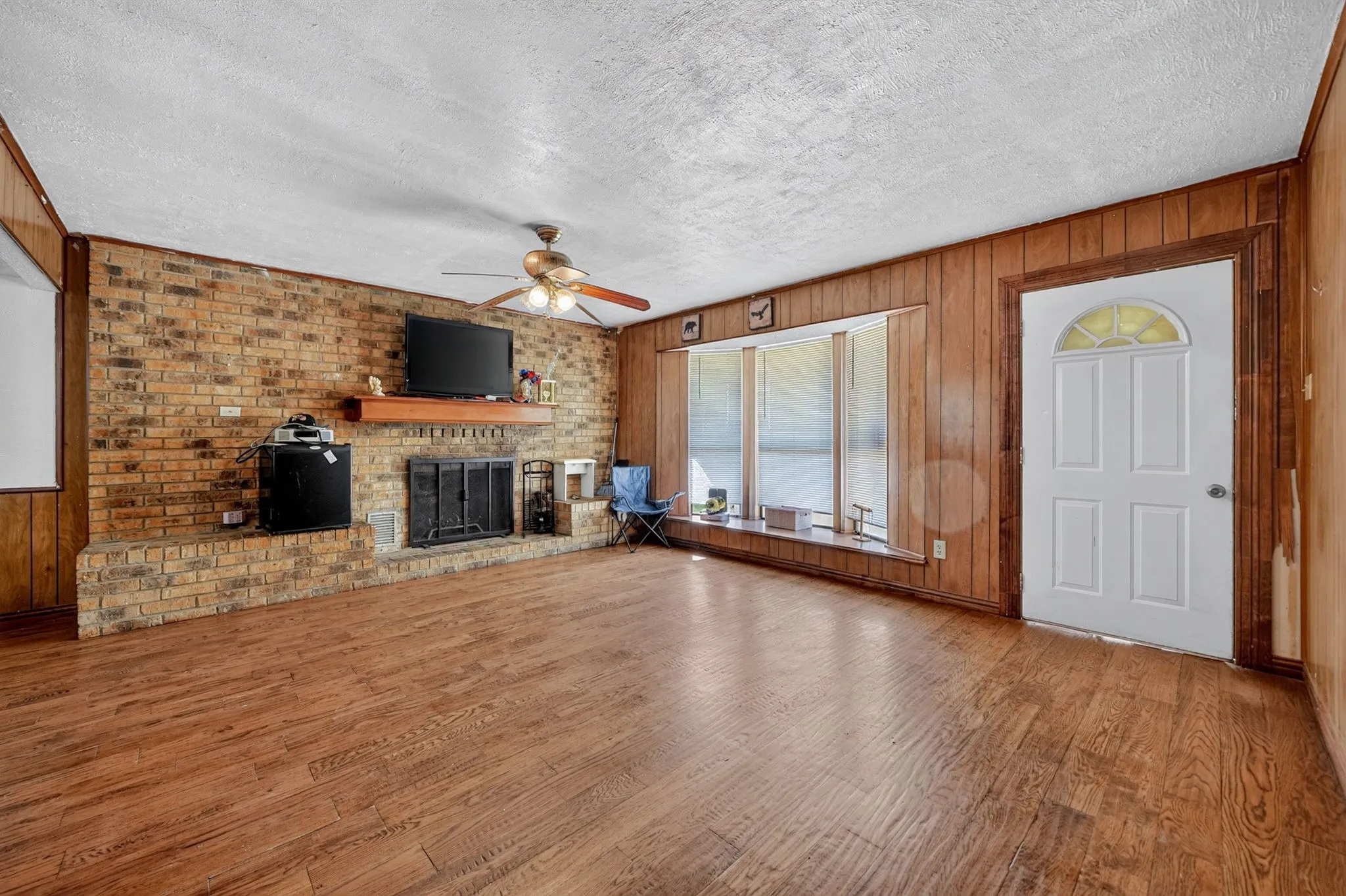 Wood-finish flooring throughout, brick accent wall with fireplace, wood paneling, bay window, and white paneled entry door with an arched glass insert
