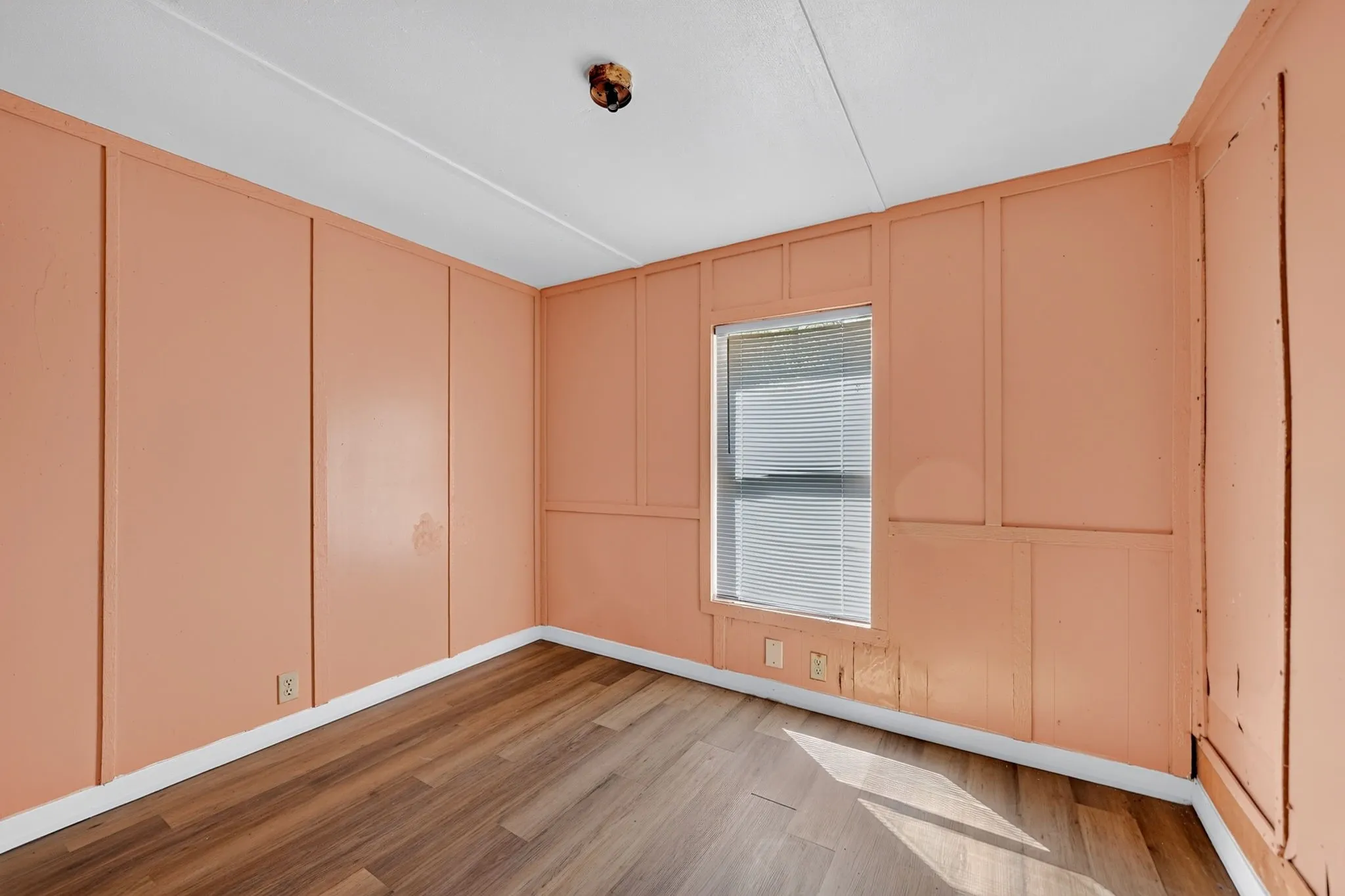 Wood-finish flooring with white trim, a single window with blinds, and paneling on the walls