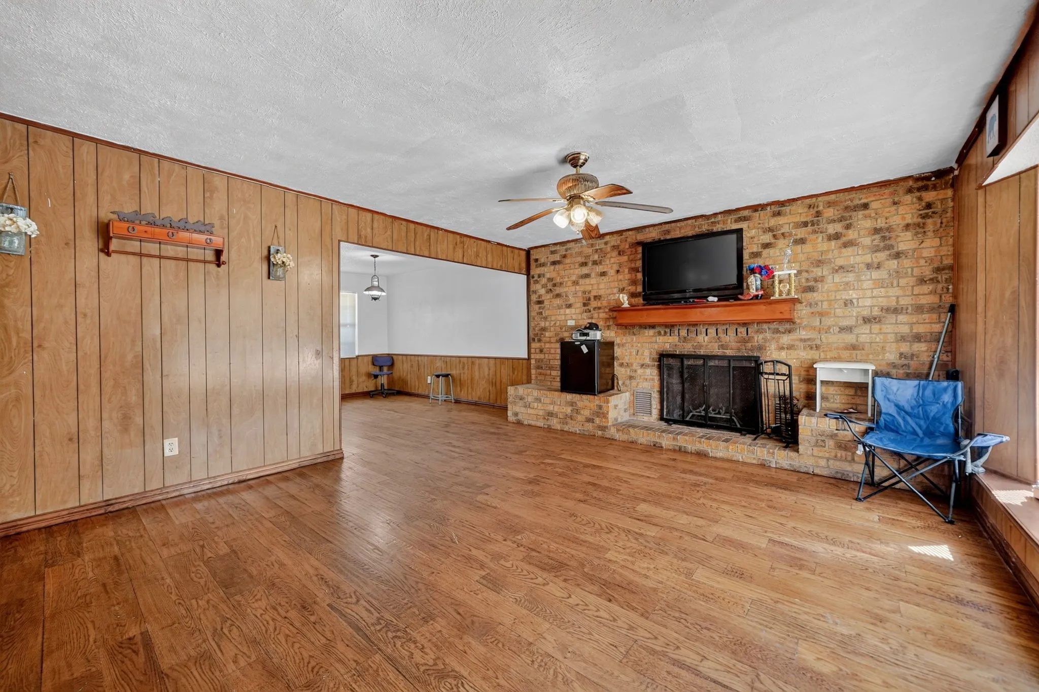 Wood-finish flooring, brick-faced fireplace with hearth, wood-paneled walls, and a ceiling fan with integrated lighting