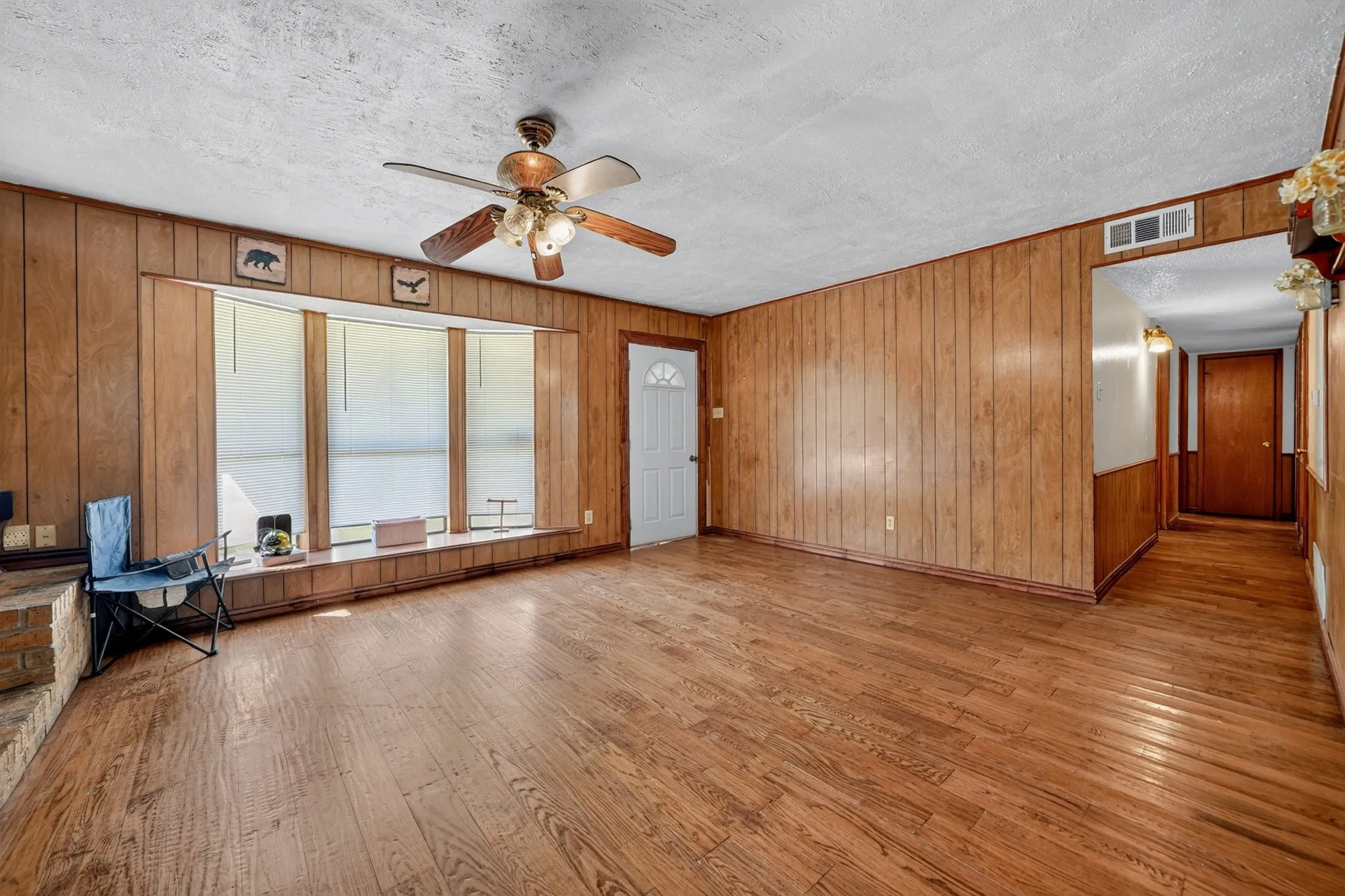 Spacious room featuring wood-finish flooring, wood paneling, a multi-pane bay window, and a ceiling fan with integrated lighting
