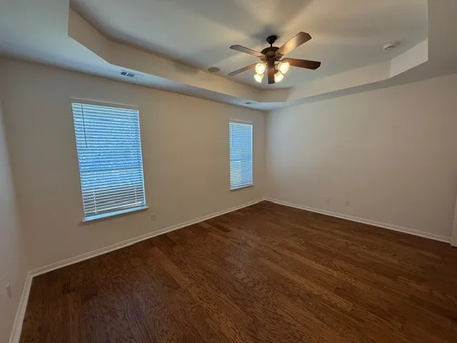 Tray ceiling with a ceiling fan, two windows with blinds, and wood-finish flooring