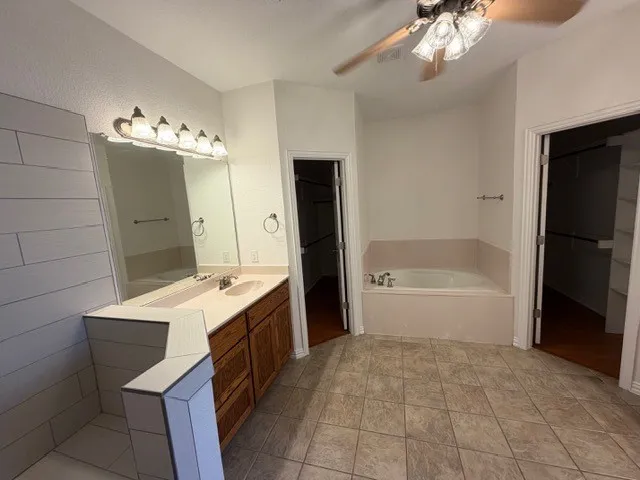 Dual vanity with wood-finish cabinetry, built-in soaking tub, tile flooring, and ceiling fan