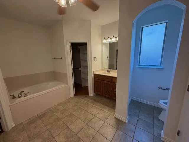 Bathroom featuring a built-in soaking tub, wood-finish vanity with countertop, wall-mounted lighting, and a separate toilet alcove