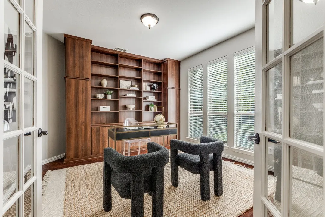 Wood-finish built-in shelving, wood-finish flooring, three vertical windows with blinds, and white French doors with frosted glass panels