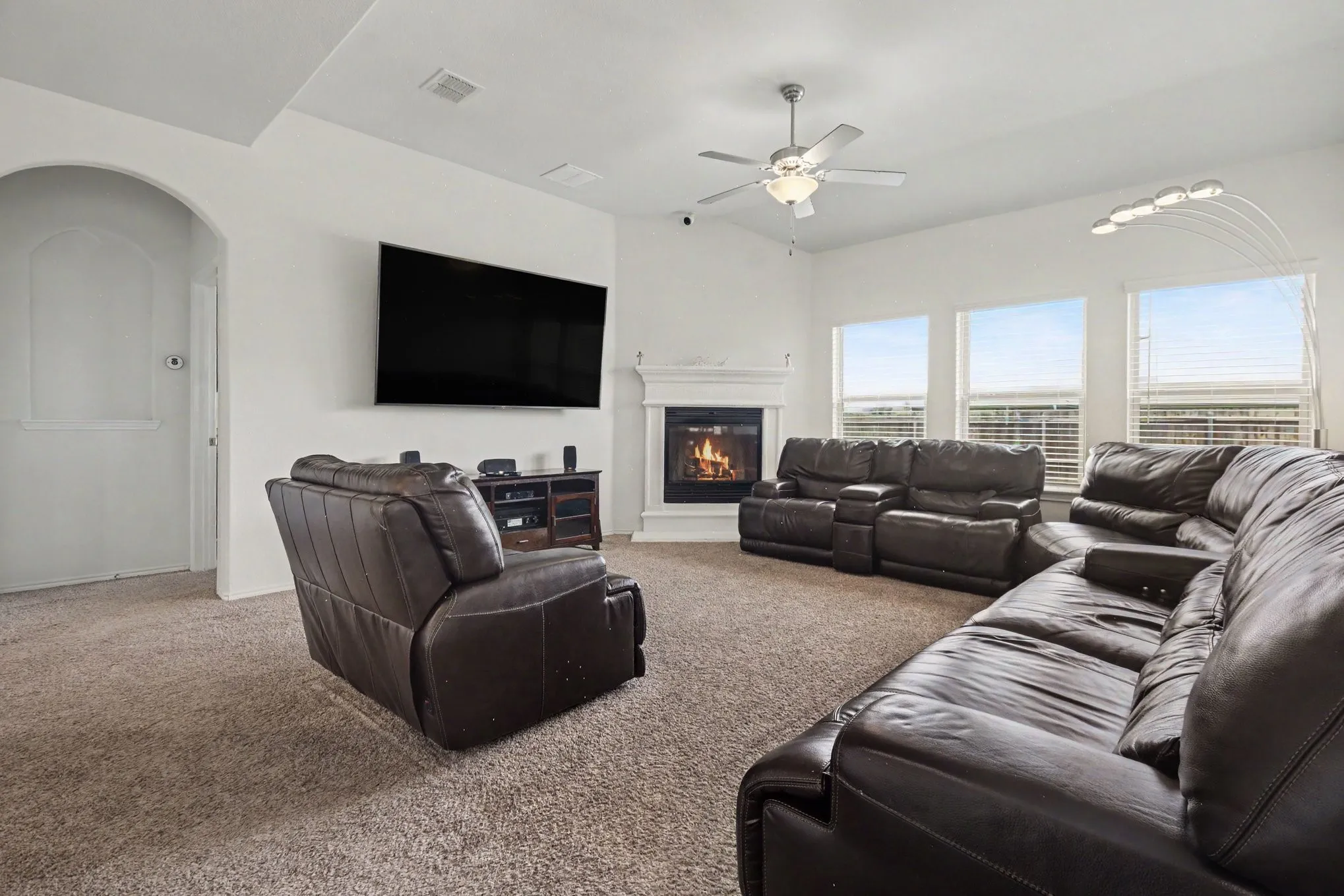 Living area featuring a corner fireplace with a white mantel, textured carpeting, multiple windows, an arched doorway, and a ceiling fan