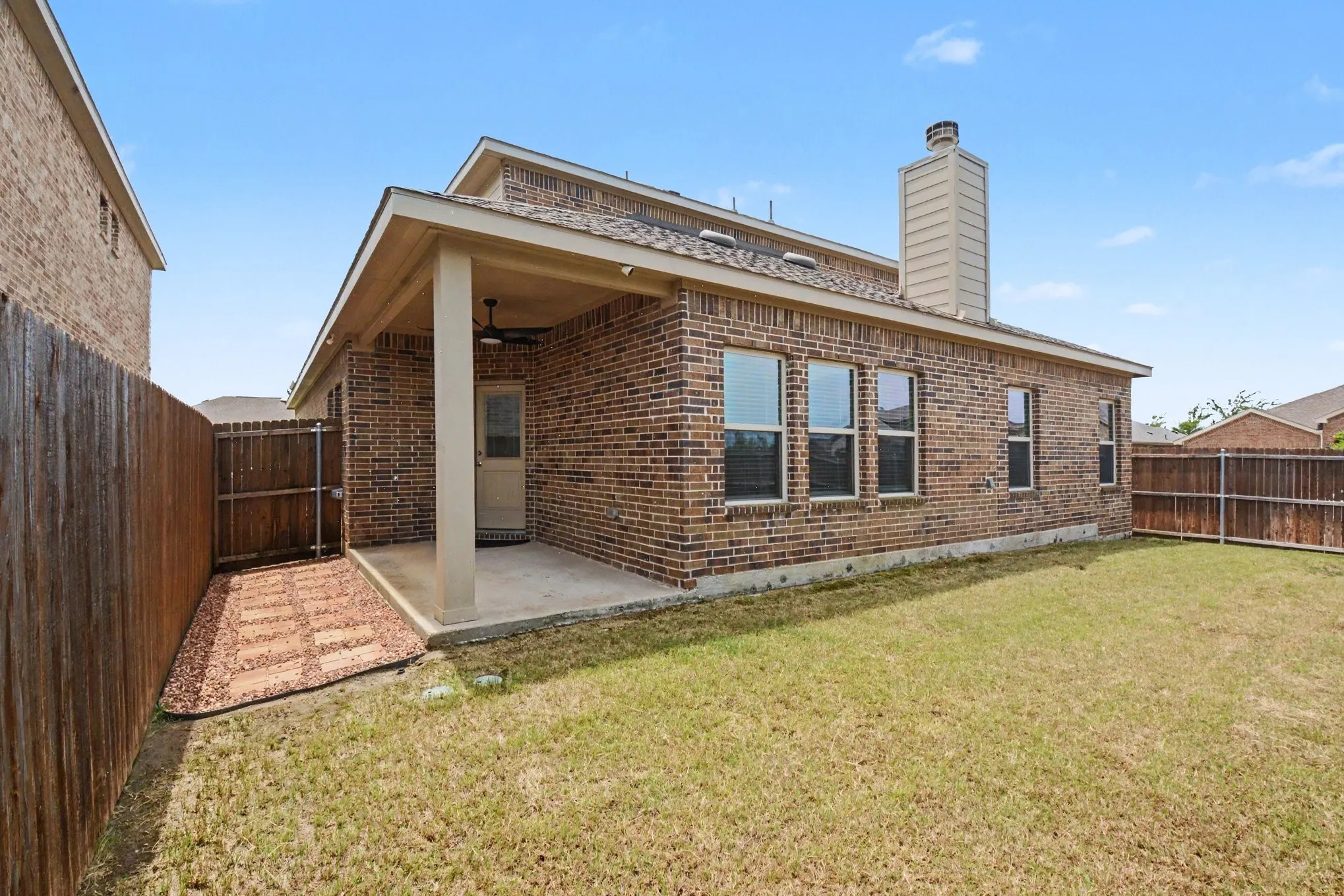Brick exterior featuring a covered patio with an overhead fan, multiple windows, a prominent chimney, and a privacy fence