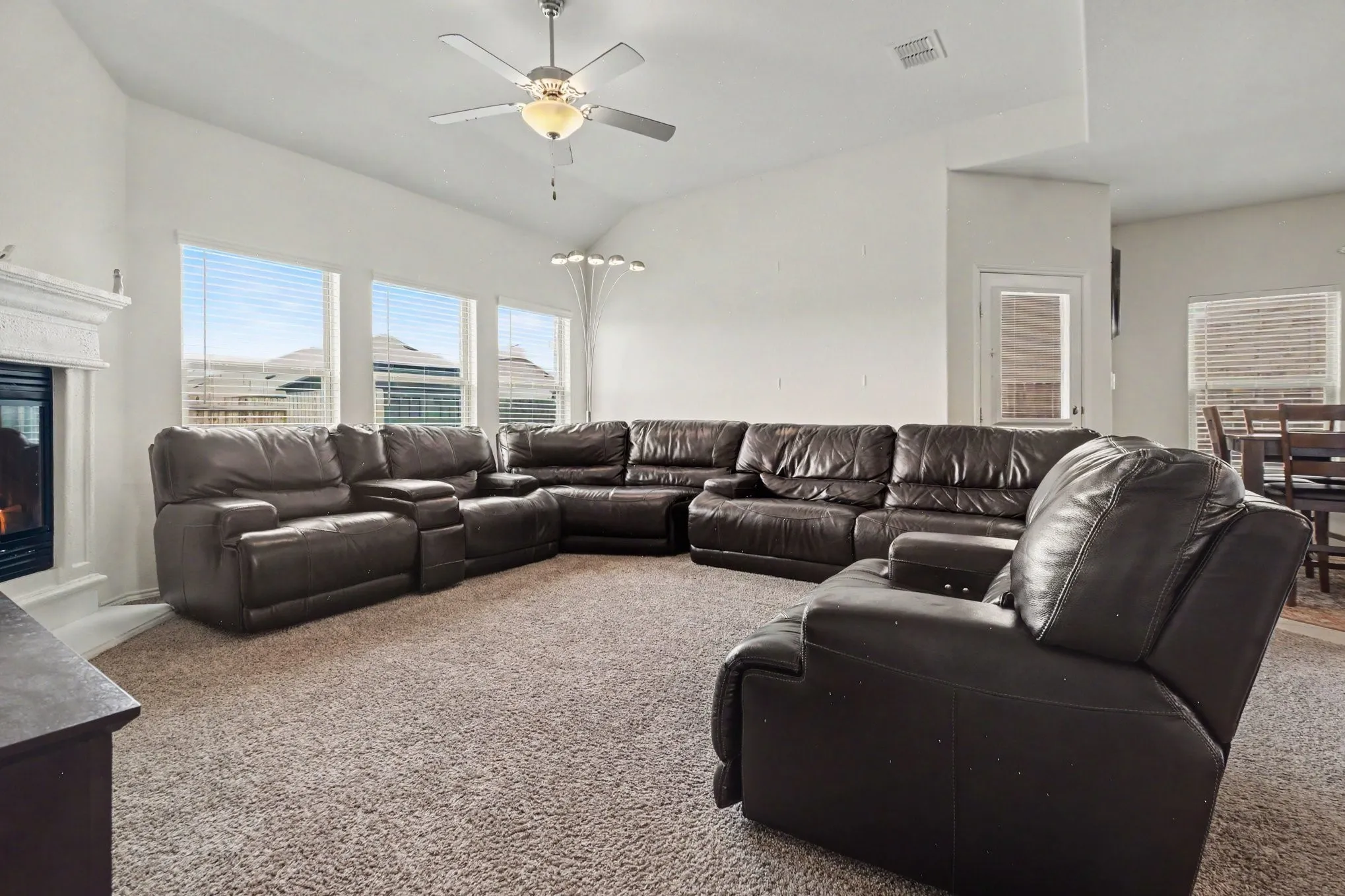 Spacious living area featuring a vaulted ceiling with a ceiling fan, a fireplace with a decorative mantel, and multiple windows with blinds