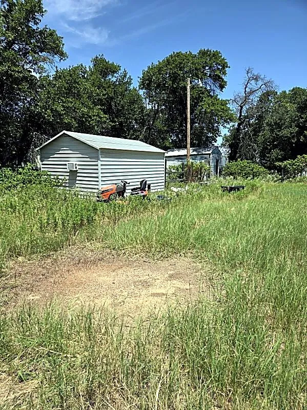 Expansive lot featuring a metal utility building with a window-mounted unit, additional rear outbuilding, and mature trees providing natural screening