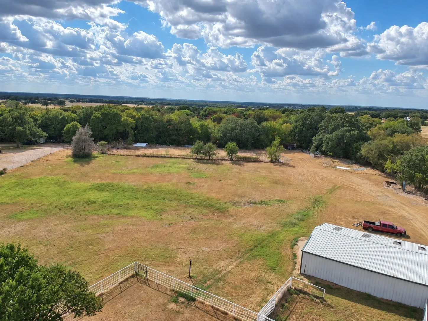 Expansive land with a metal outbuilding, fenced enclosures, and a tree-lined perimeter