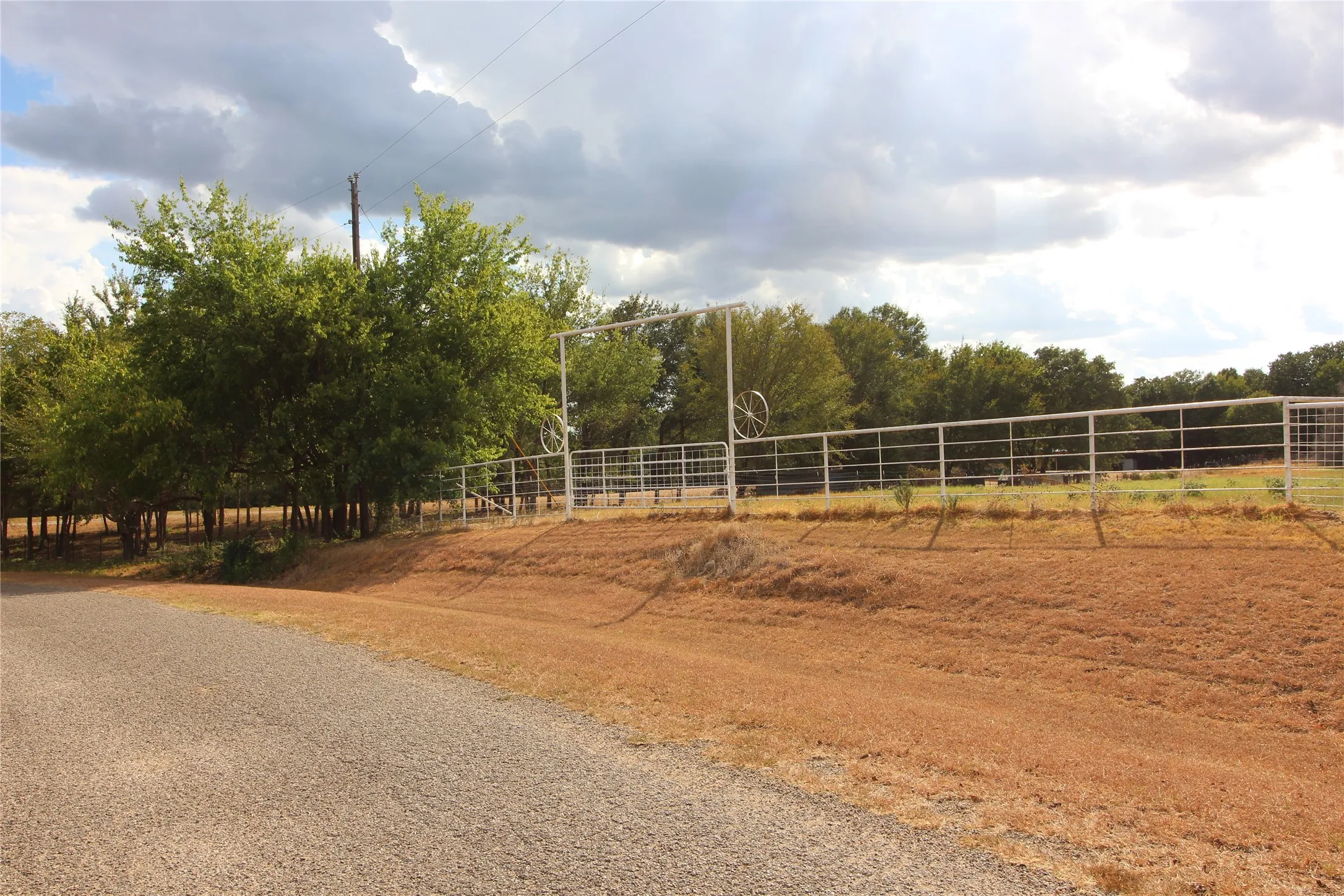 Gated entrance with white metal fencing, a gravel driveway, and mature trees lining the property