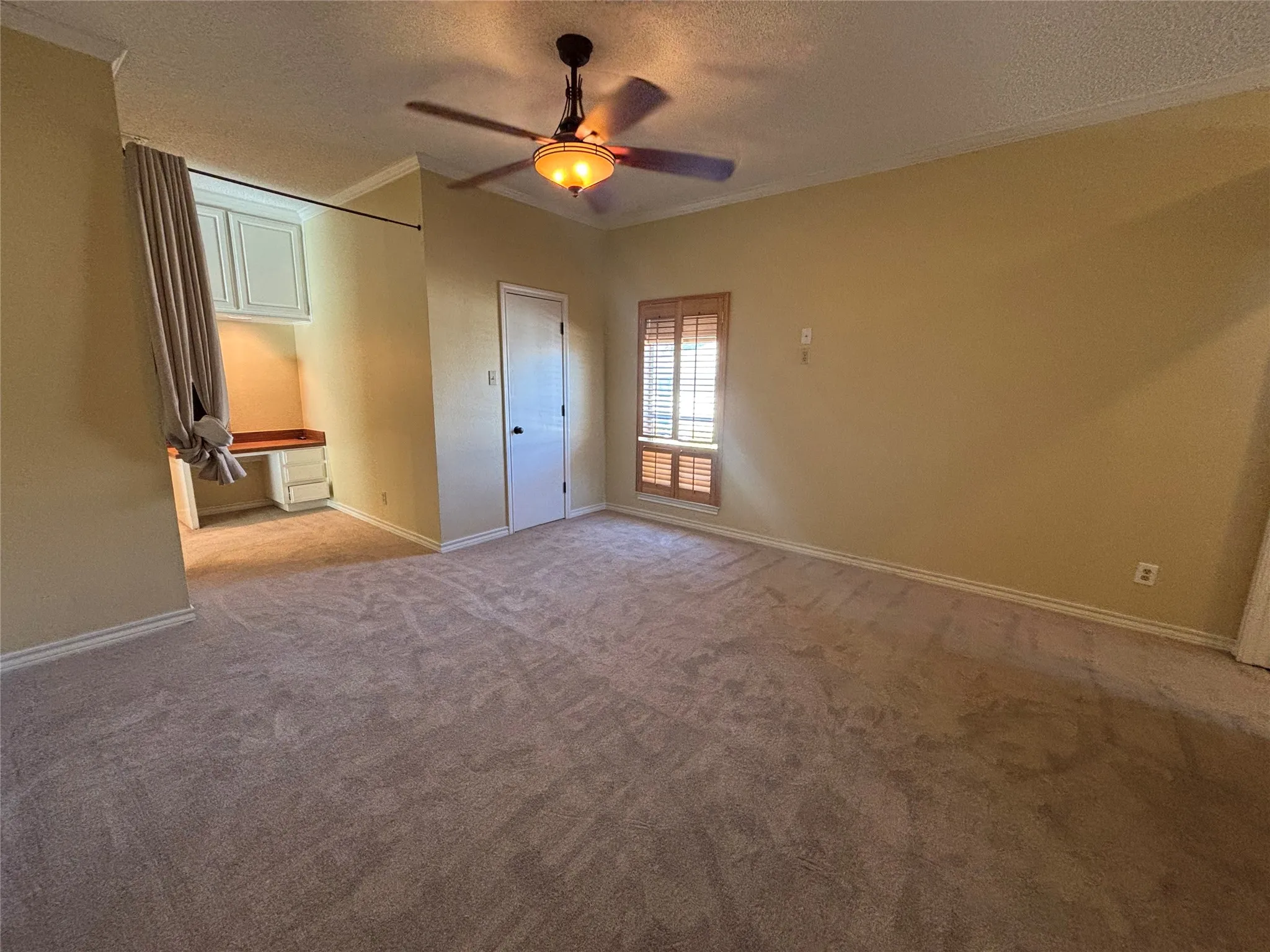 Spacious room with neutral carpeting, built-in desk with cabinetry, window with wood blinds, and ceiling fan