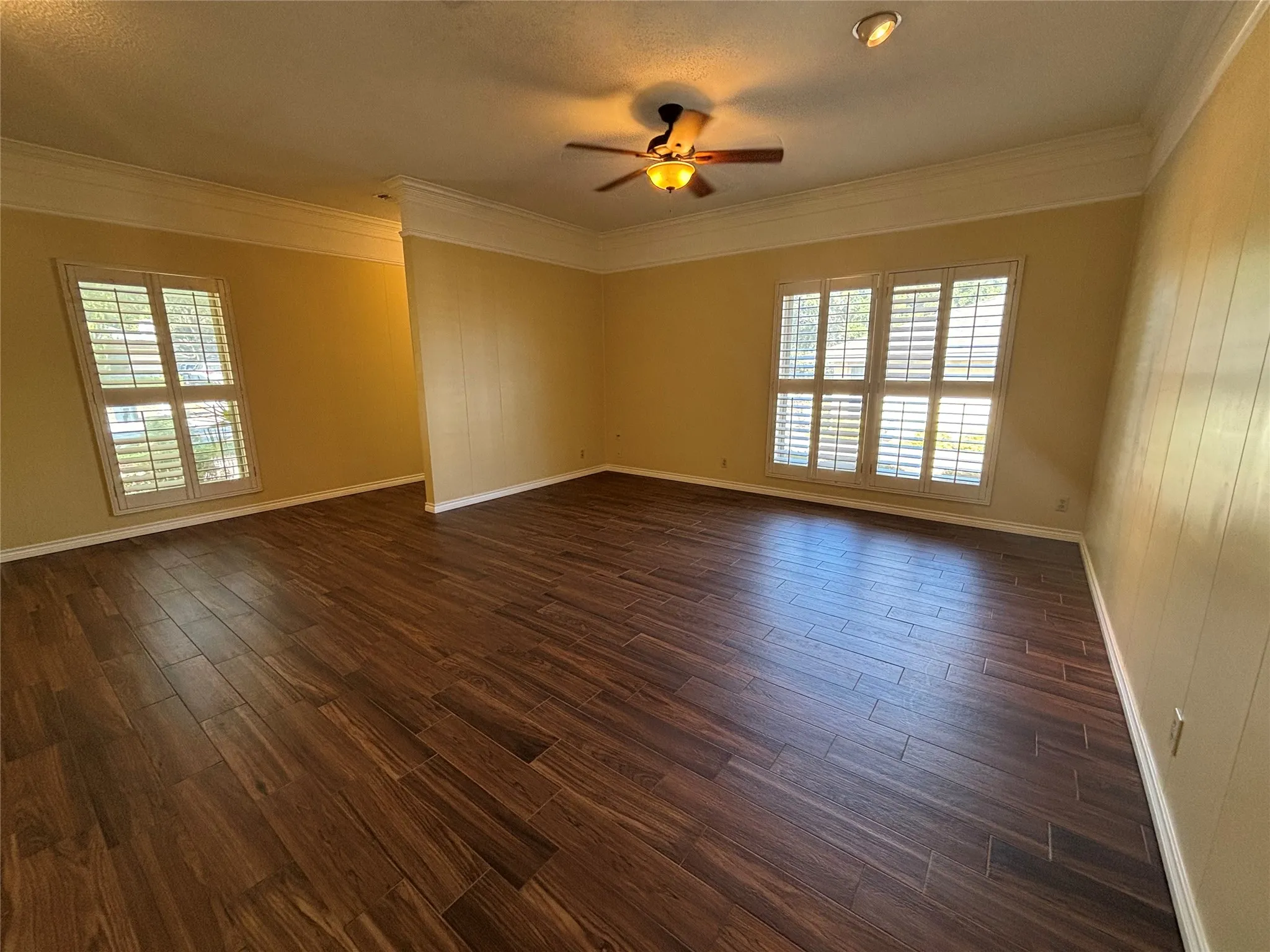Wood-finish flooring, two windows with plantation shutters, crown molding, and a ceiling fan with integrated lighting