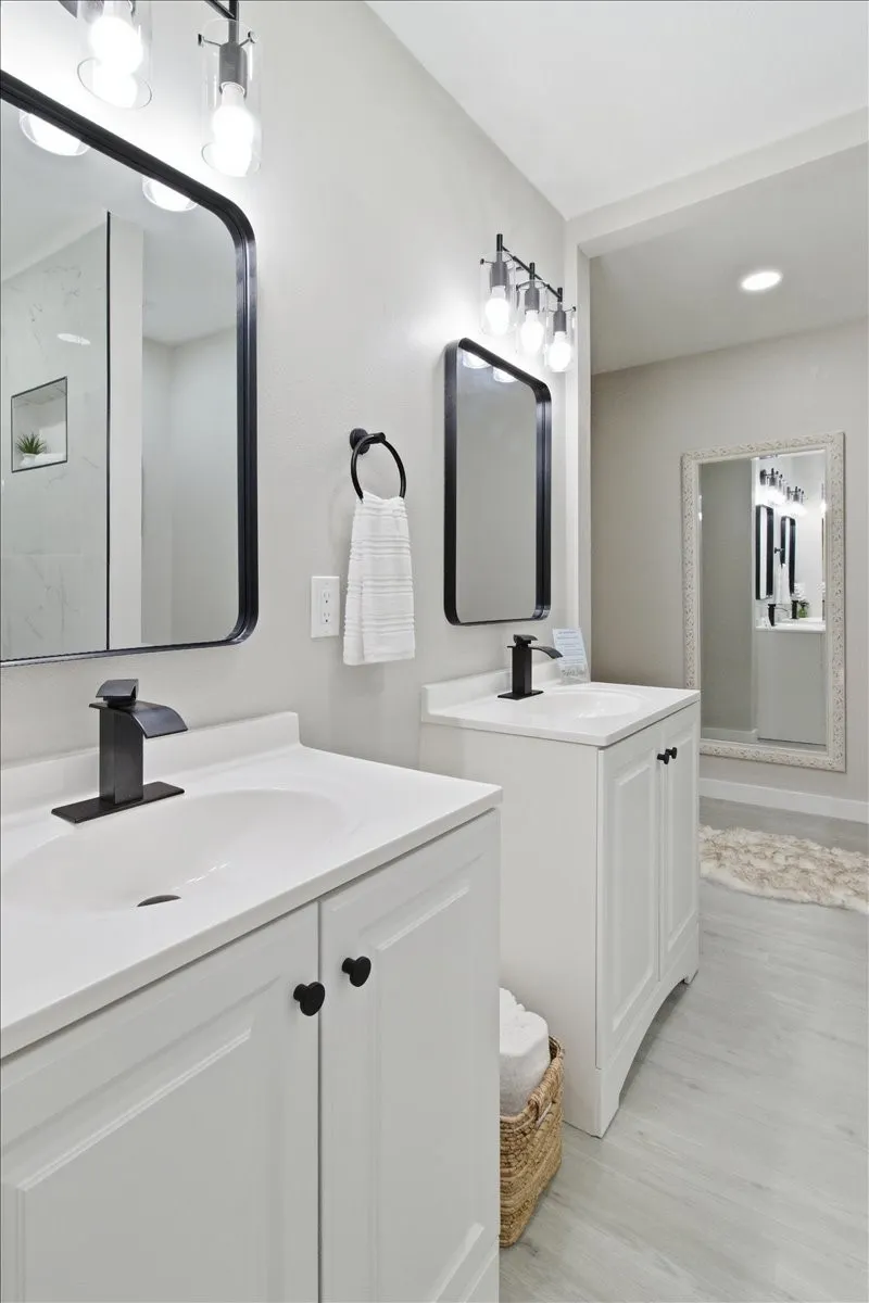 Dual white vanities with integrated sinks and matte black fixtures, framed mirrors, and overhead lighting