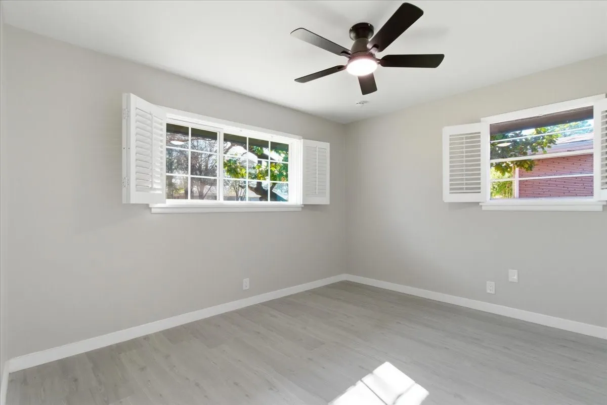 Light gray walls, wood-finish flooring, white window shutters, two large windows, and a ceiling fan with integrated lighting