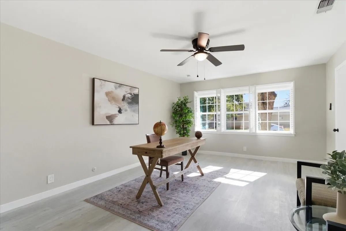 Bright room with light wood-finish flooring, three large windows, a ceiling fan with integrated lighting, and neutral wall tones