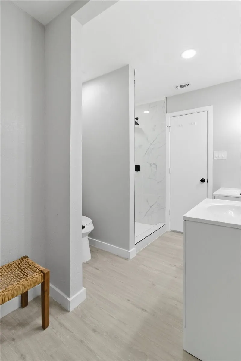 Light wood-finish flooring, recessed lighting, glass-enclosed shower with tiled walls, white vanity with integrated sink, and a white toilet