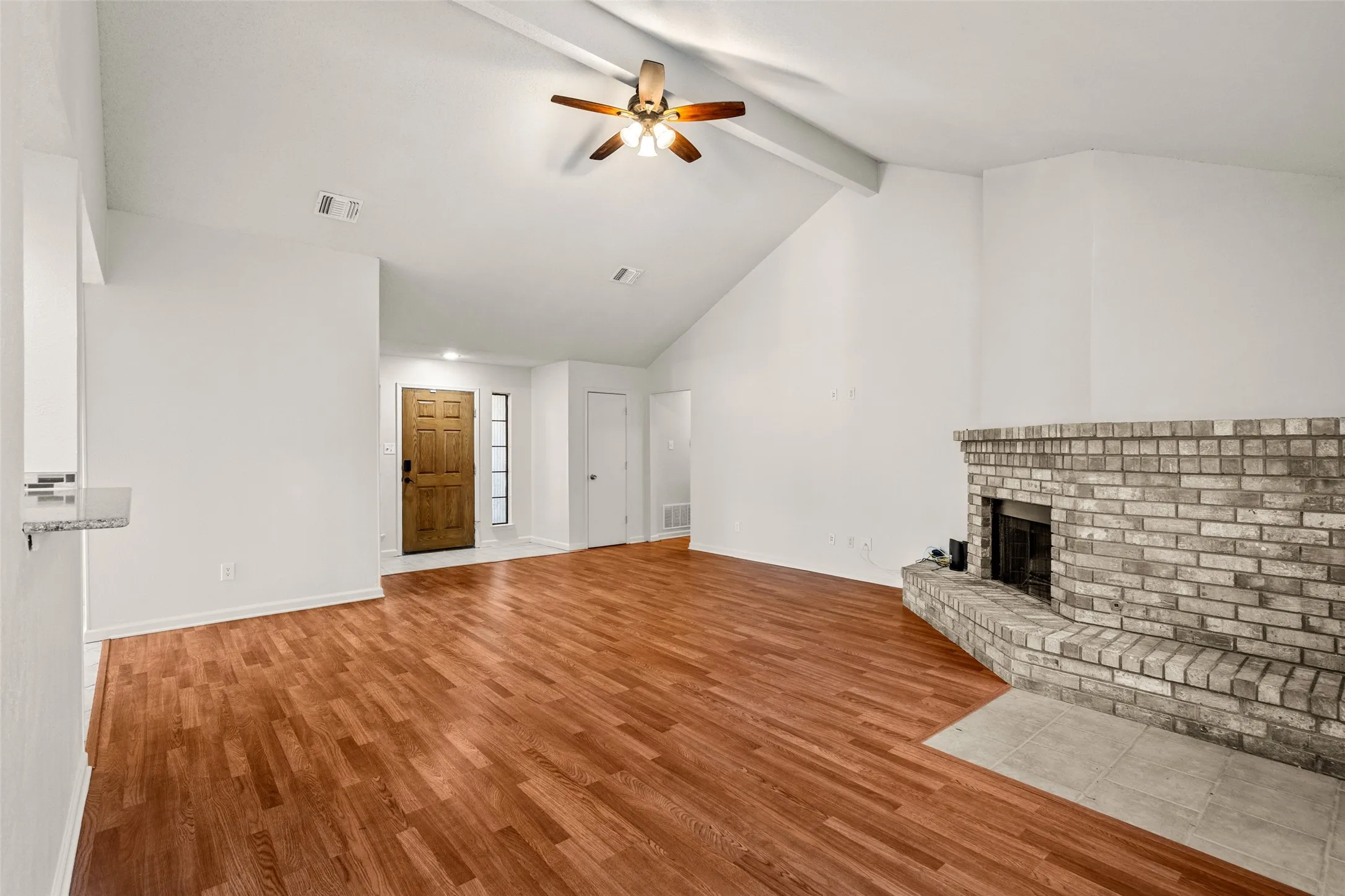 Vaulted ceilings with exposed beam, wood-finish flooring, brick fireplace with hearth, ceiling fan, and a paneled wood entry door with sidelight
