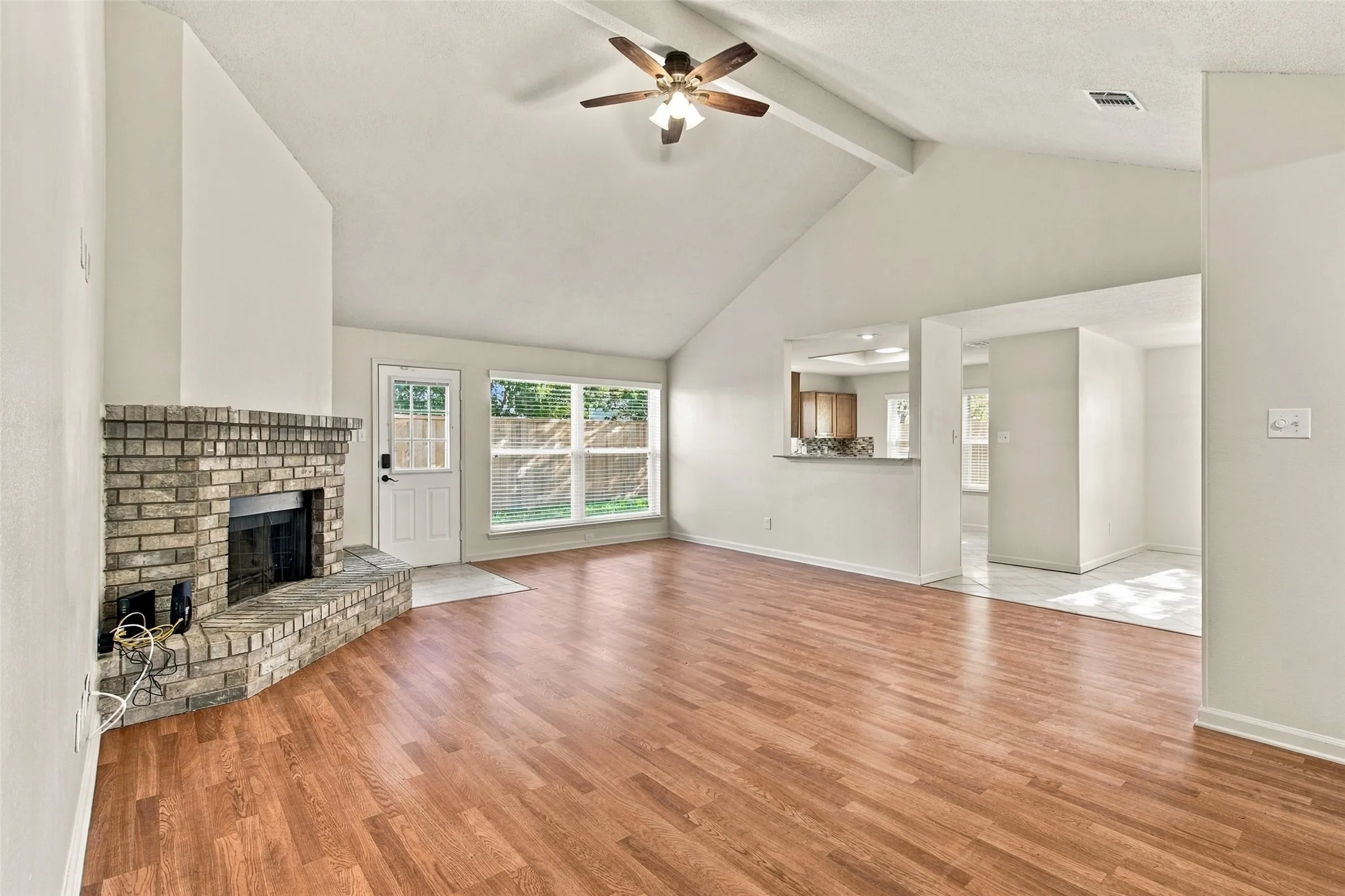 Vaulted ceiling with exposed beam, wood-finish flooring, brick fireplace with hearth, ceiling fan with light fixture, and pass-through window to kitchen