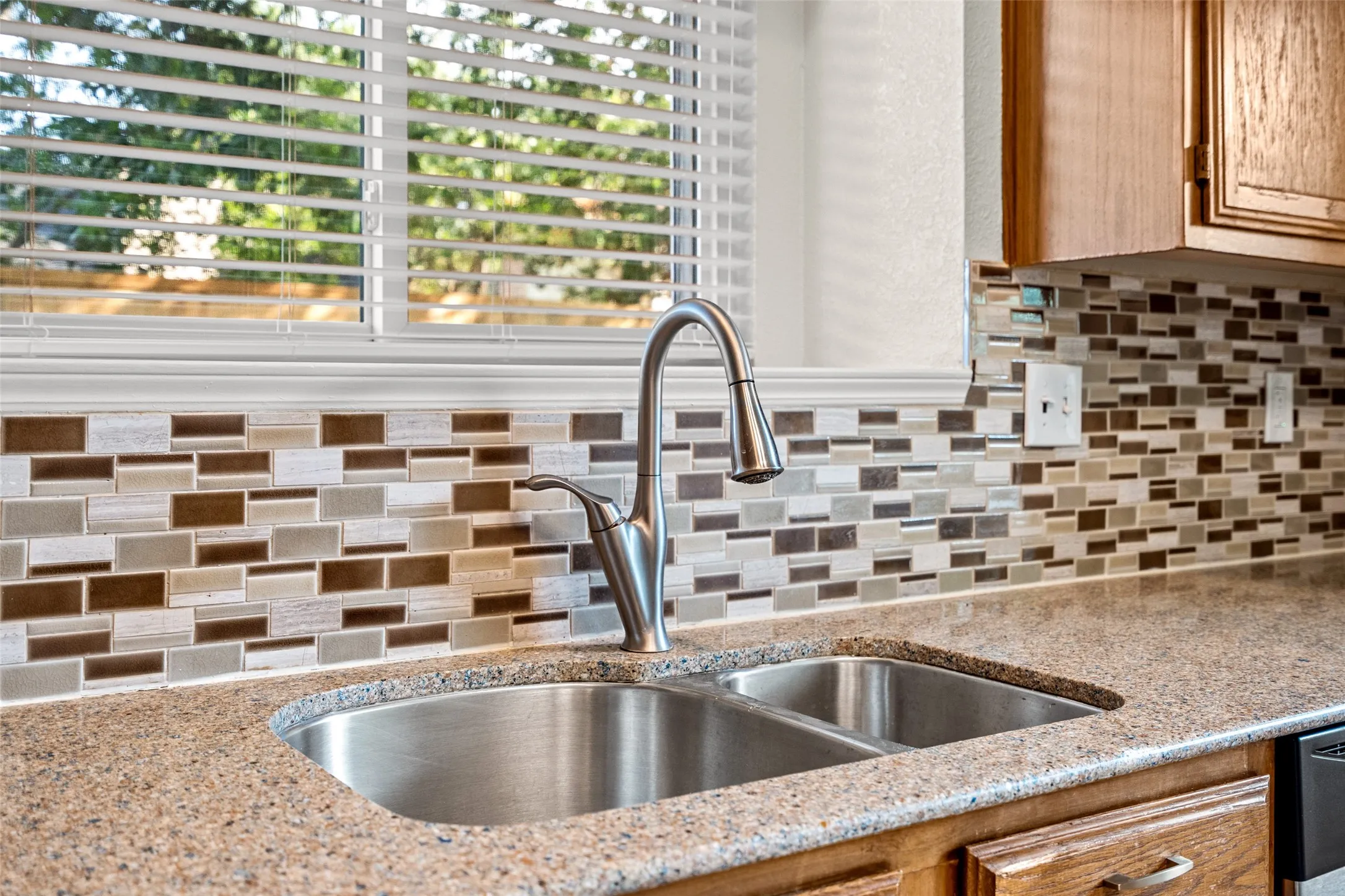 Dual basin stainless steel sink with a high-arc faucet, complemented by a multi-toned mosaic tile backsplash and speckled countertop