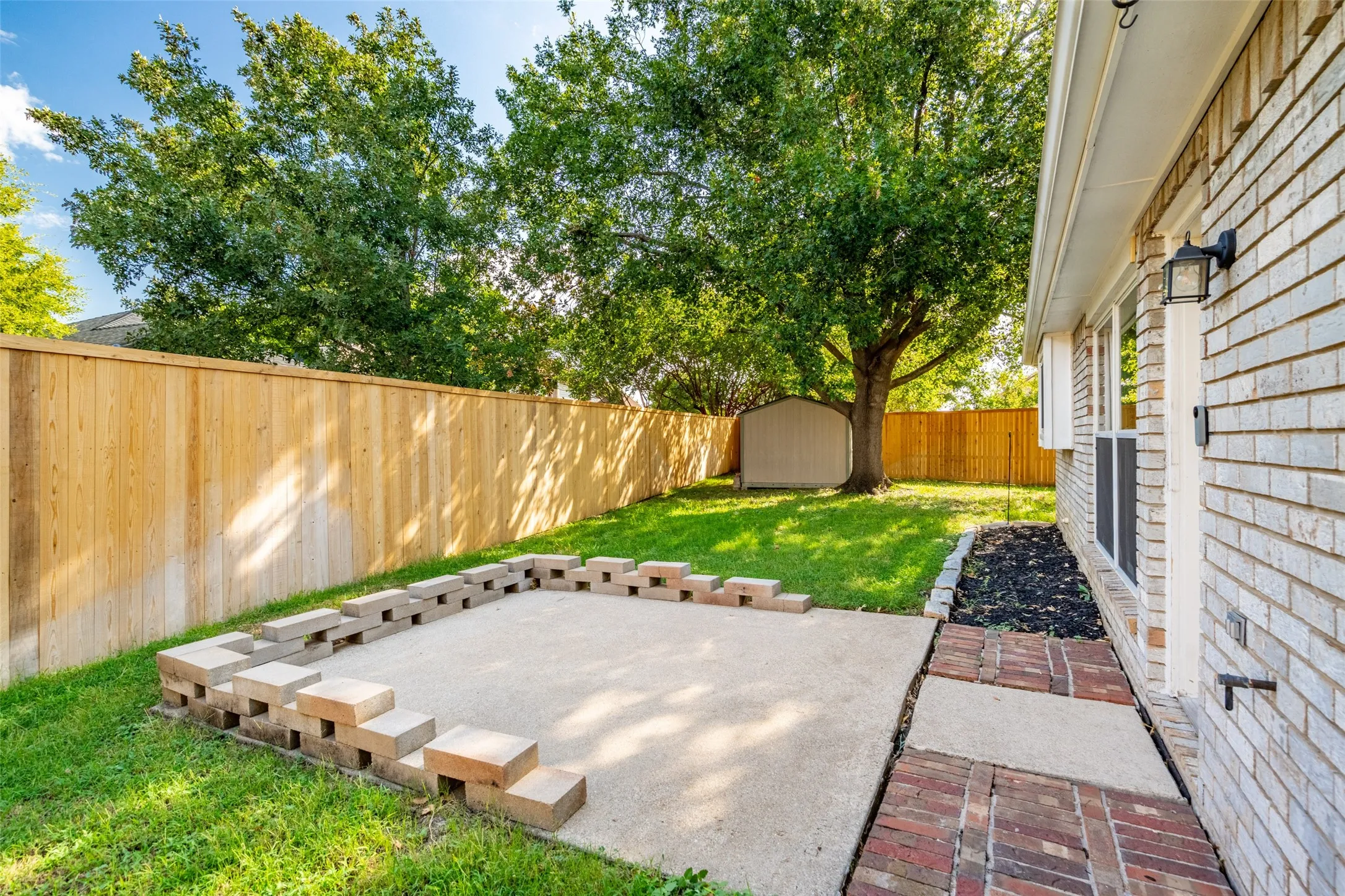 Private fenced backyard featuring a concrete patio with block retaining wall, storage shed, and a brick exterior