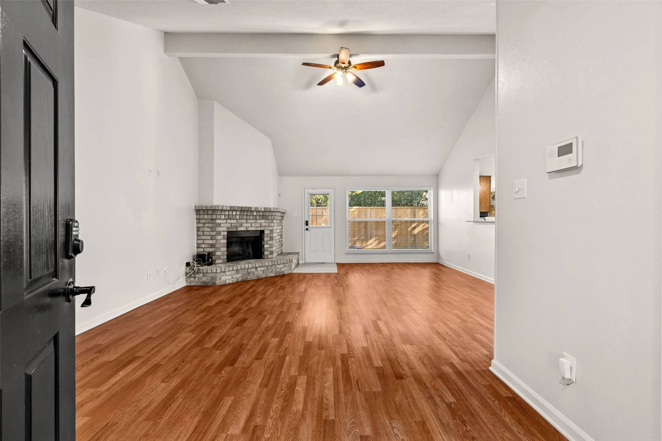 Vaulted ceiling with exposed beam, wood-finish flooring, brick fireplace, ceiling fan, and large windows