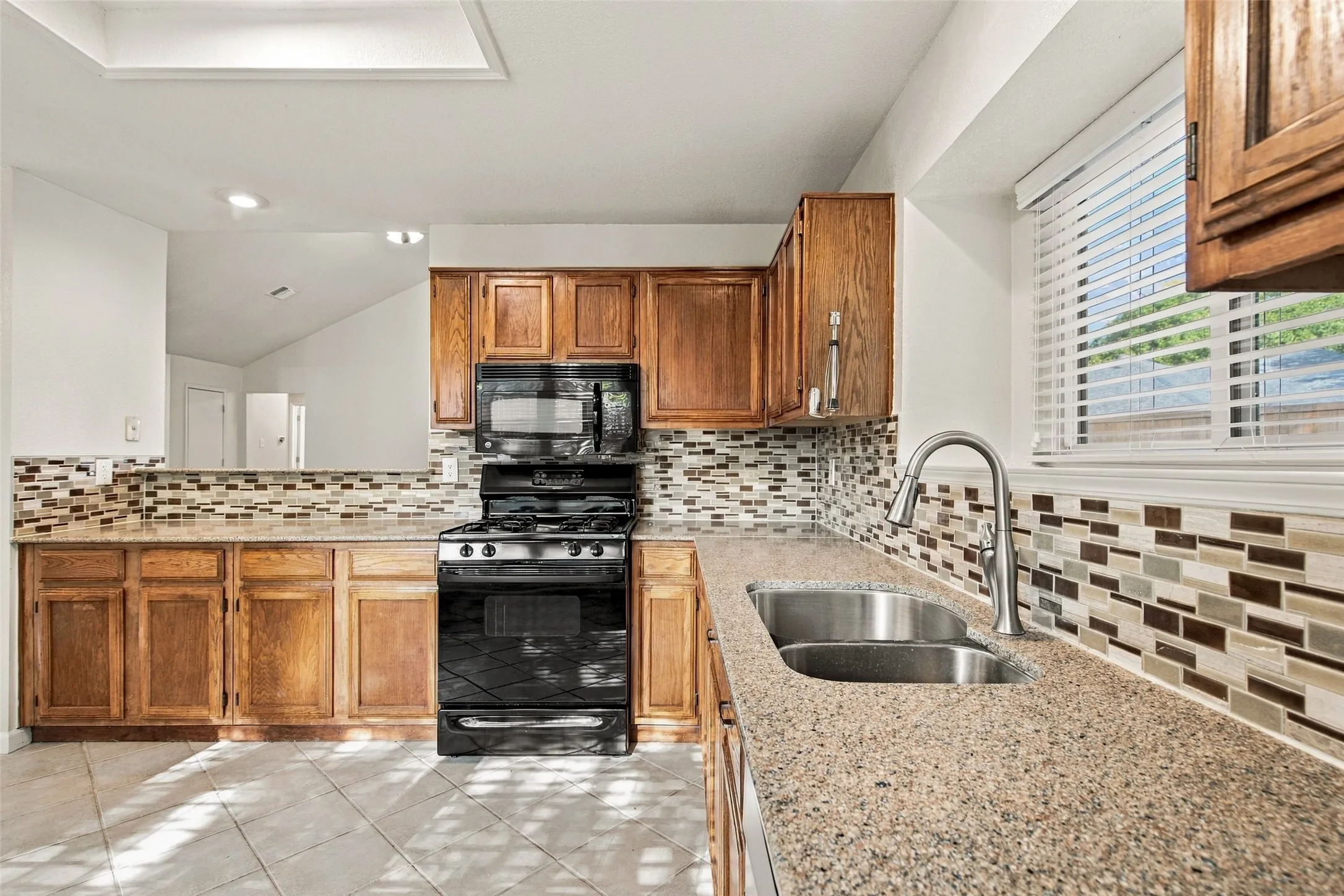 Granite-finish countertops, mosaic tile backsplash, wood cabinetry, stainless steel double basin sink, and a black appliance package