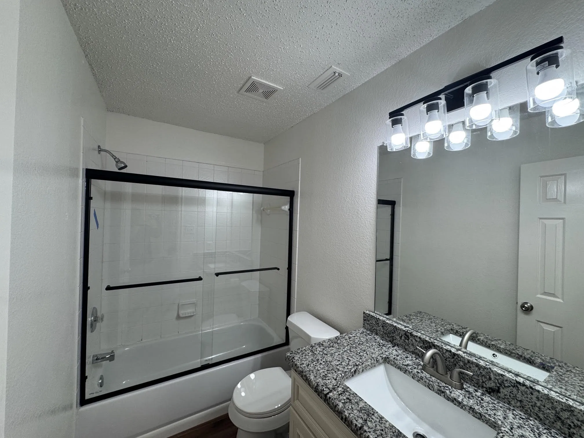 Modernized bathroom featuring a full tub with sliding glass doors, a granite vanity countertop, and updated overhead lighting
