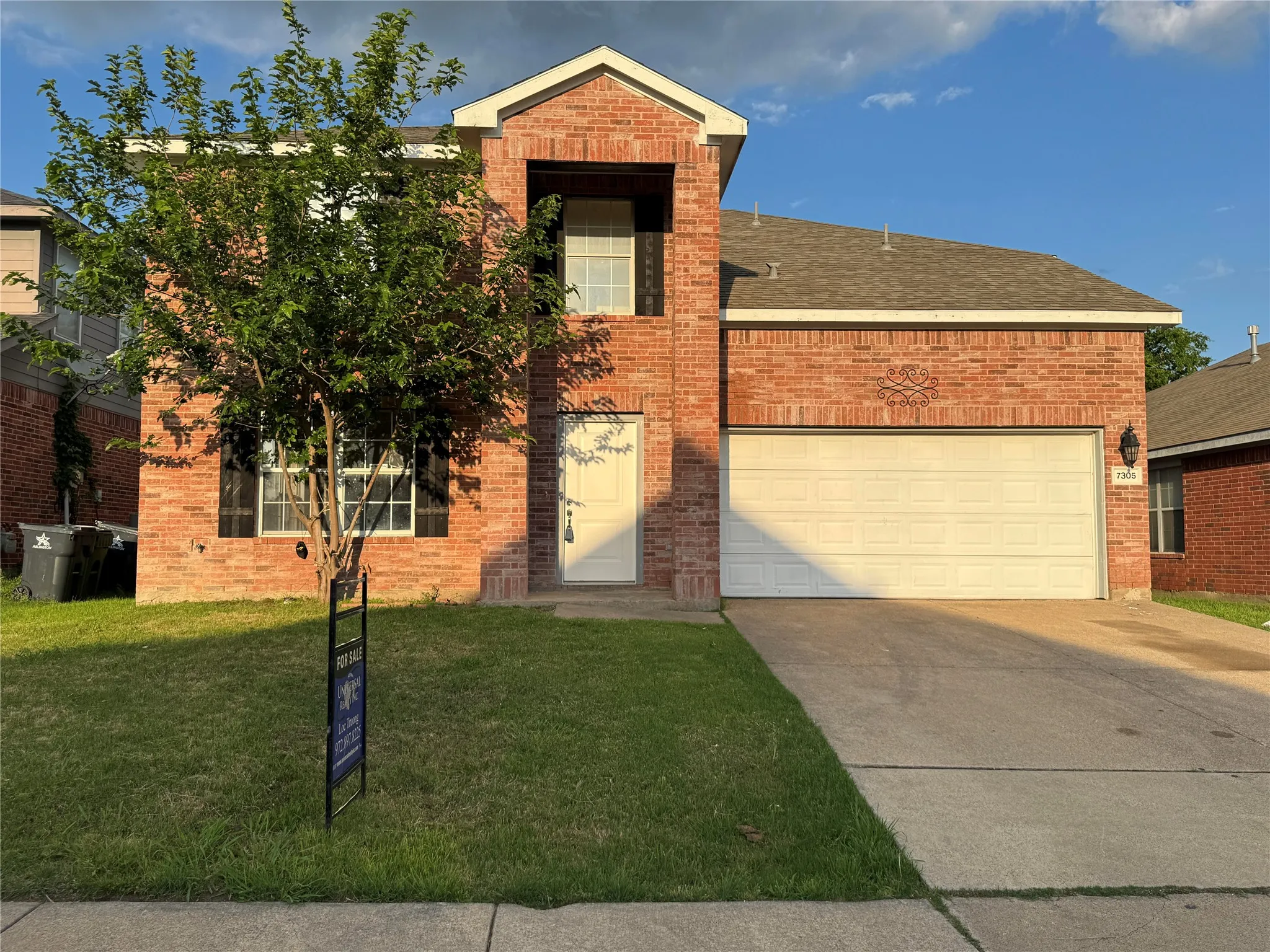 Traditional brick exterior featuring a prominent two-car garage, manicured lawn, and mature front tree