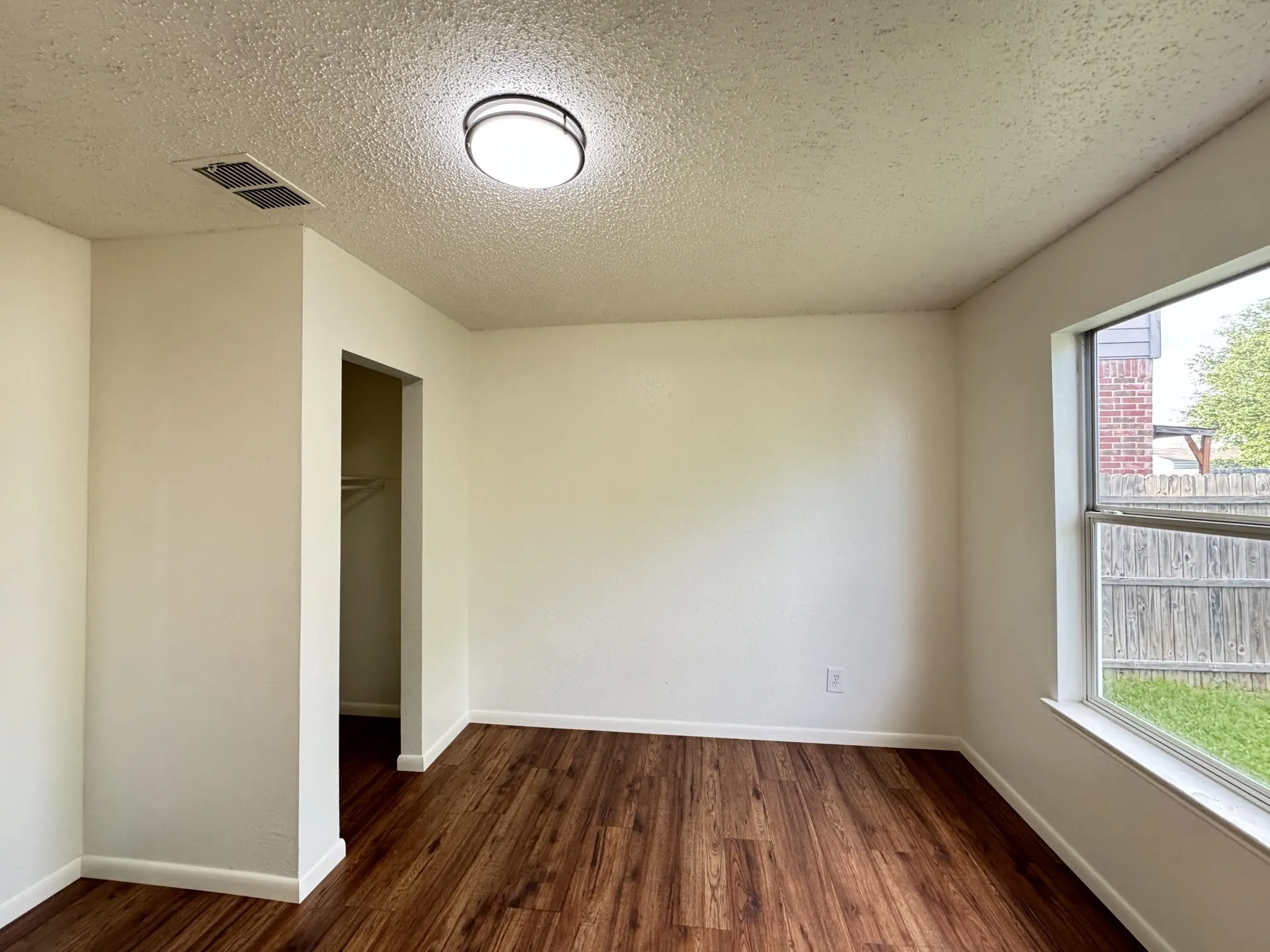 Spacious interior room featuring wood-finish flooring, white baseboards, and a window with a backyard outlook