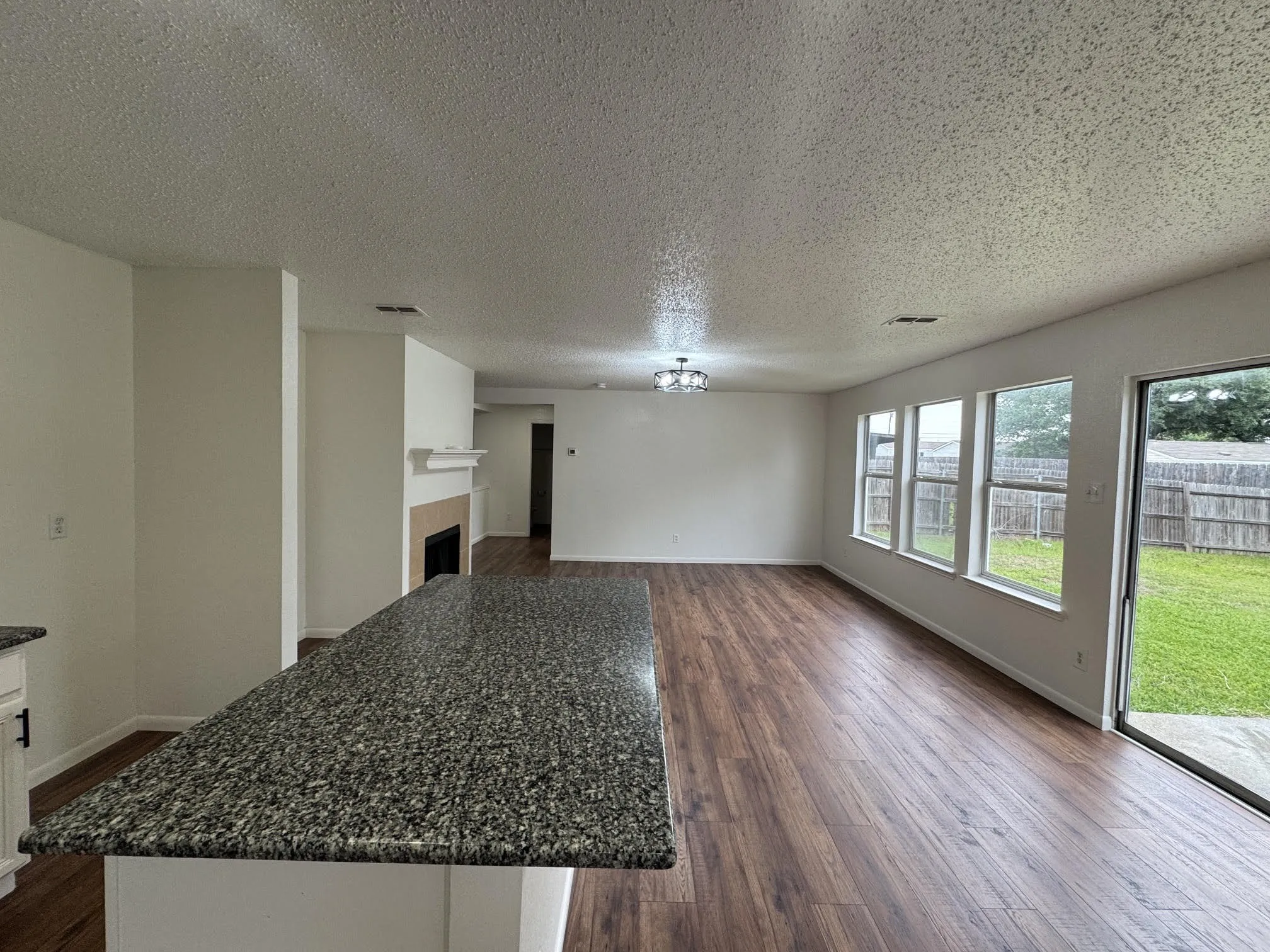 Kitchen island with a granite-style countertop, wood-finish flooring, a fireplace with mantel, multiple windows, and a sliding glass door opening to a fenced yard