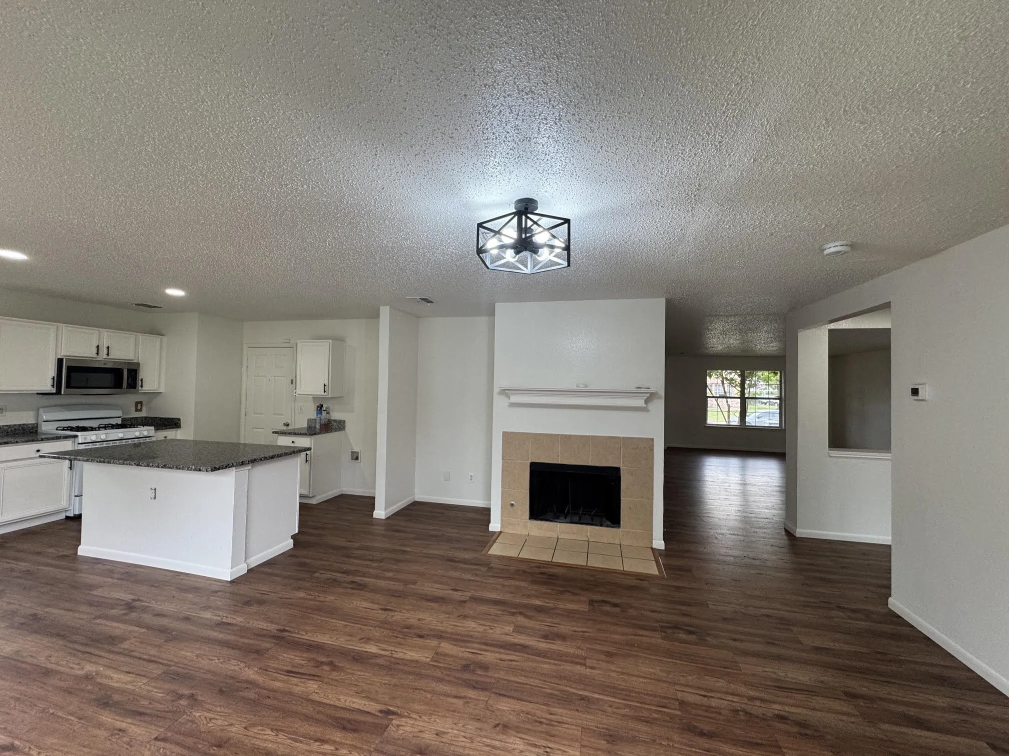 Open-concept main living space featuring wood-finish flooring, a kitchen island with granite-style countertop, white cabinetry with stainless steel appliances, and a tiled fireplace
