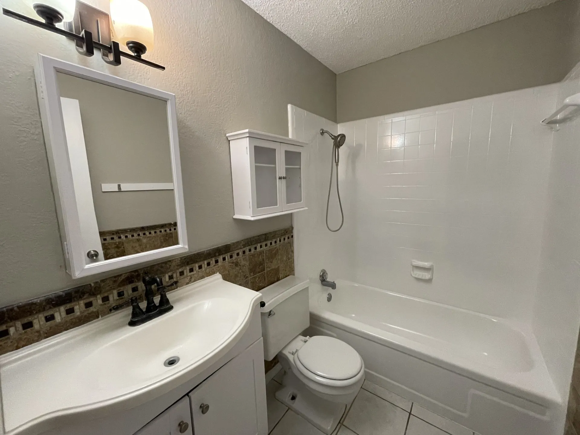 Bright bathroom featuring a curved vanity with an integrated sink, decorative tile accent band, framed mirror with integrated lighting, and a tile-surround tub/shower combination with glass enclosure and wall-mounted showerhead