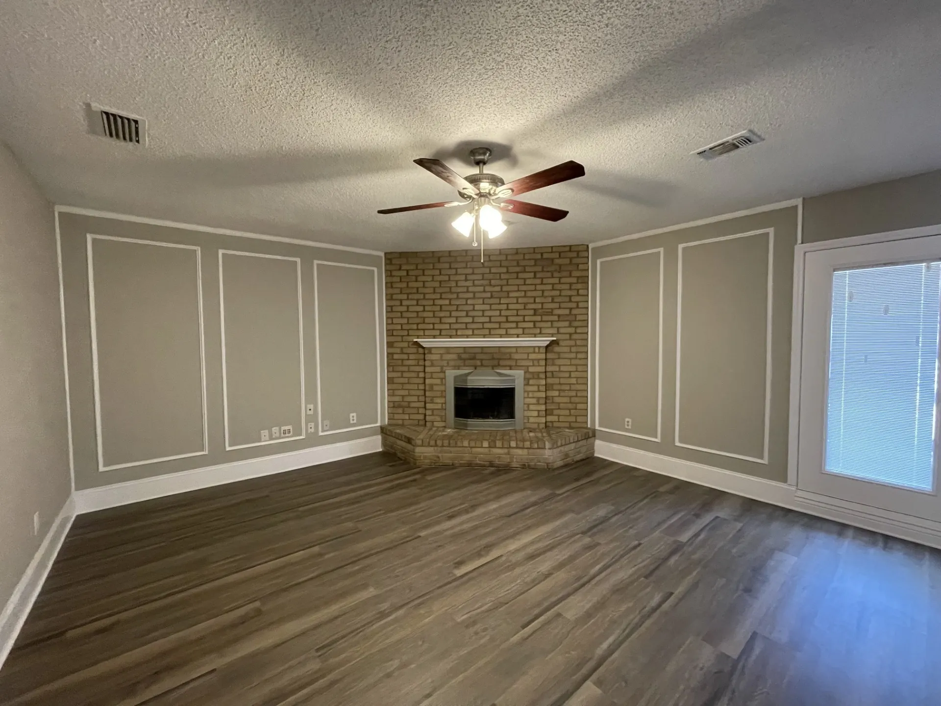 Spacious living area featuring wood-finish flooring throughout, decorative wall paneling with white trim, brick fireplace with integrated hearth, ceiling fan with integrated lighting, and glass sliding door providing natural light