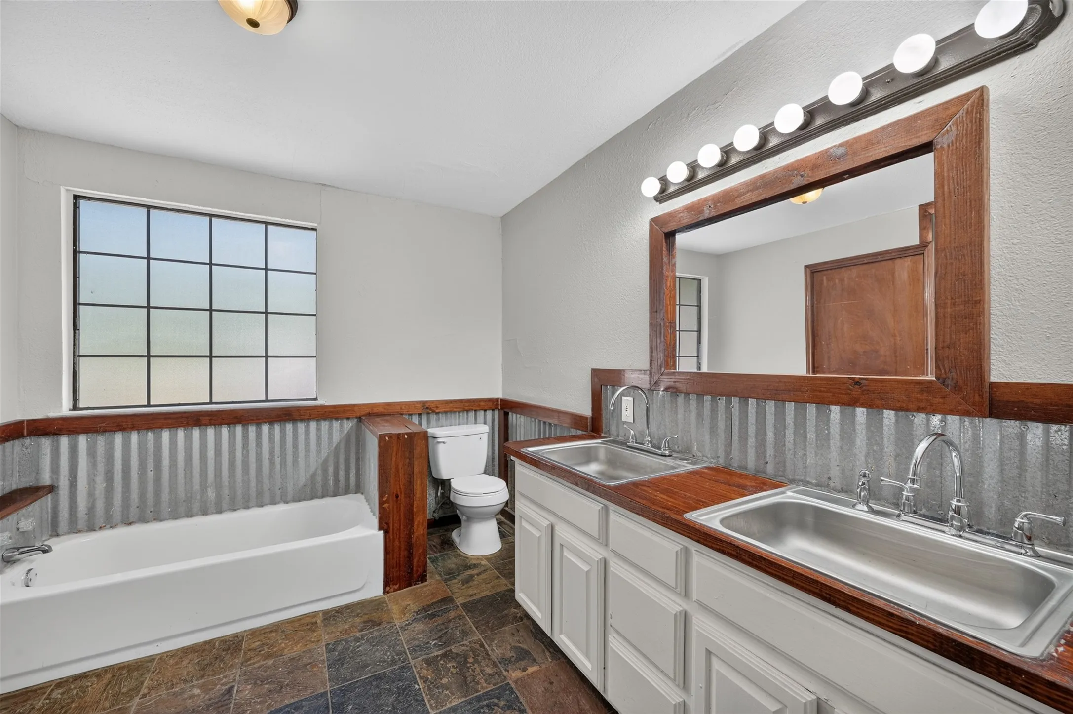Dual stainless steel sinks, wood-finish countertops, corrugated metal wainscoting, a framed mirror with vanity lighting, and a window with grid detailing